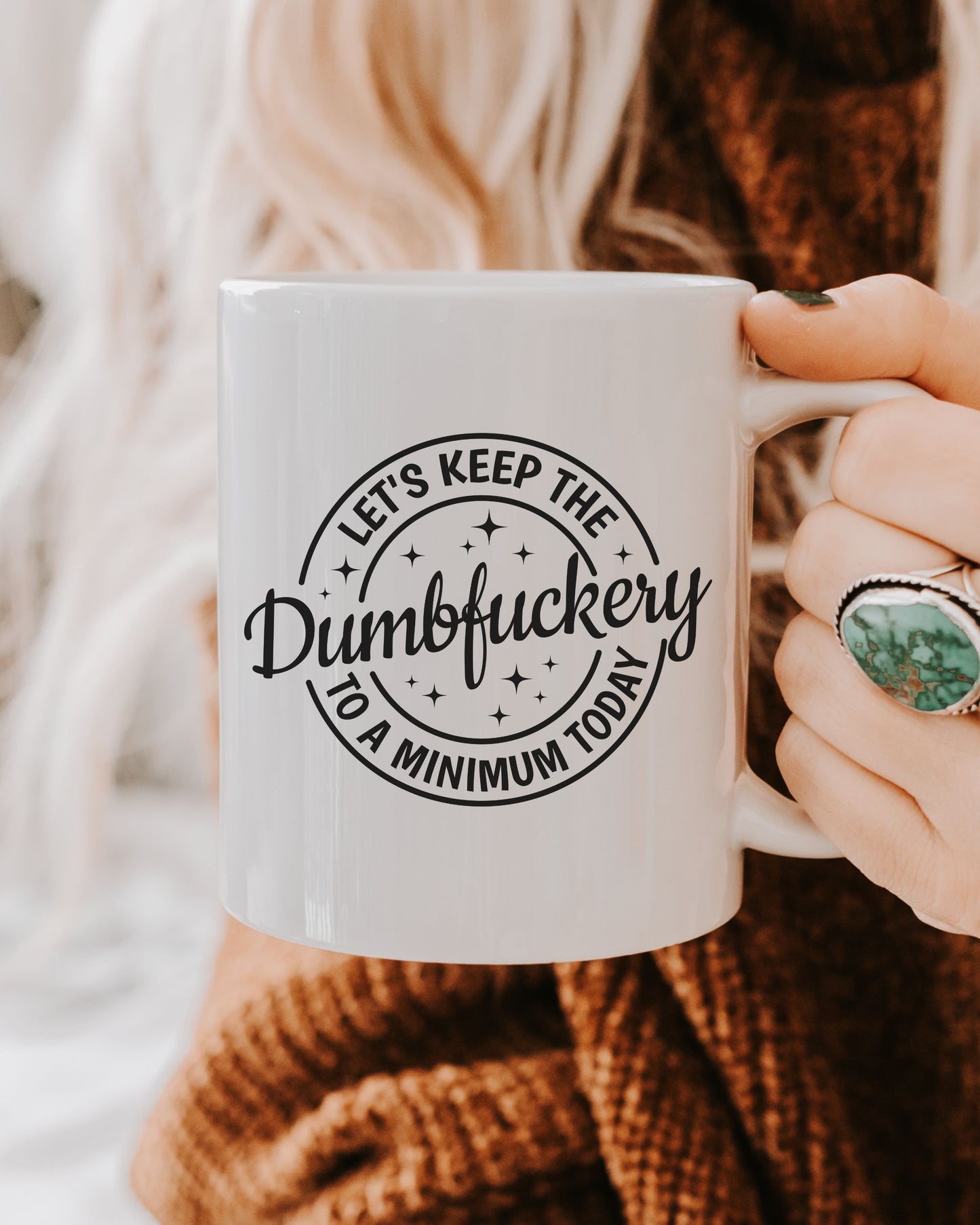 Person holding a mug with text 'Let's keep the dumbfuckery to a minimum today' against a neutral background