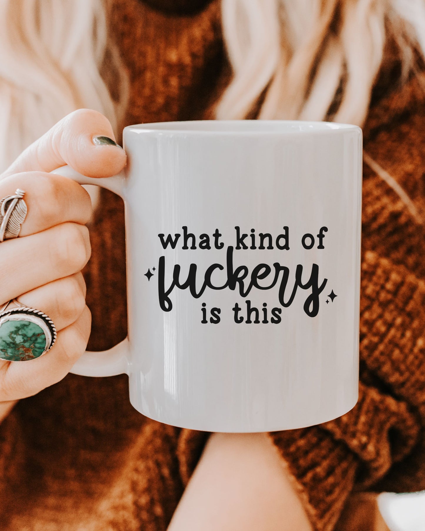 Person holding a mug with text 'what kind of fuckery is this' against a blurred background