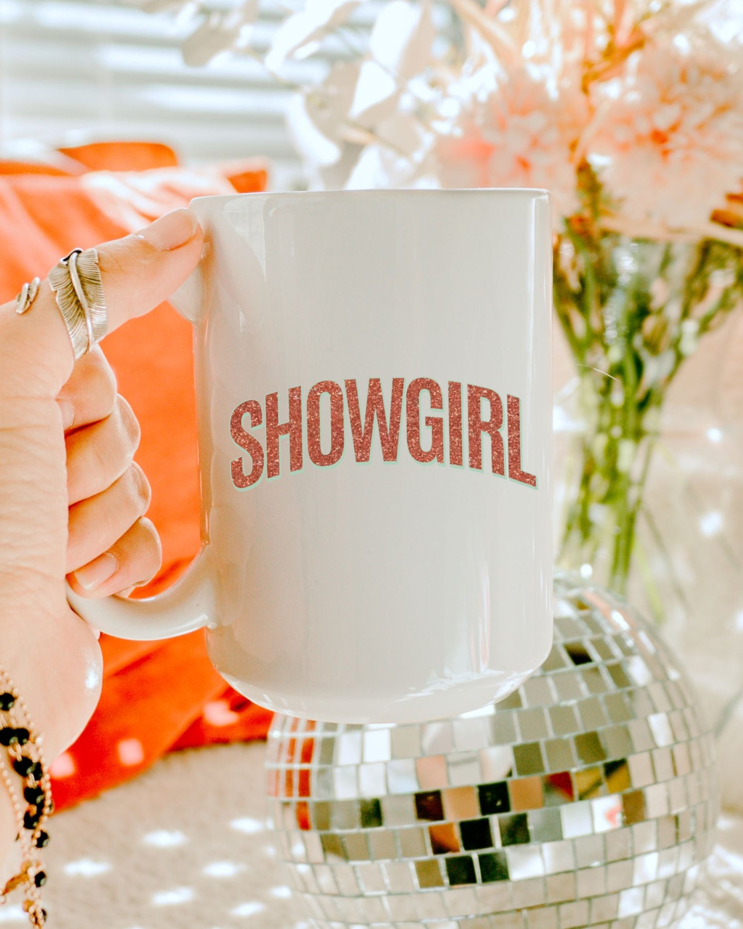 Person holding a 'SHOWGIRL' mug with a disco ball and flowers in the background