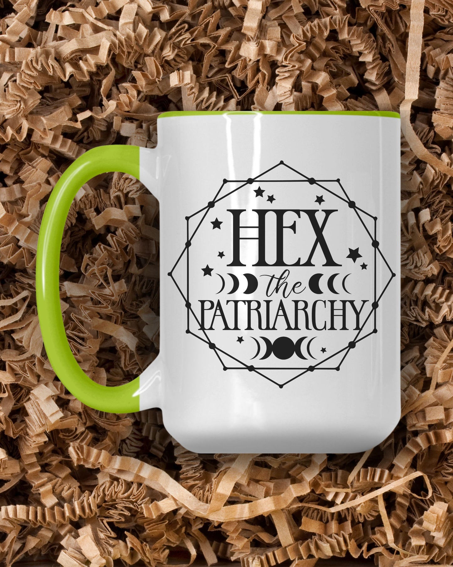 White mug with green handle featuring 'Hex the Patriarchy' design on a bed of brown shredded paper.