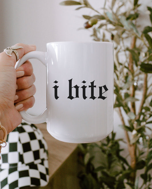White mug with 'i bite' text held by a hand, blurred plant in the background