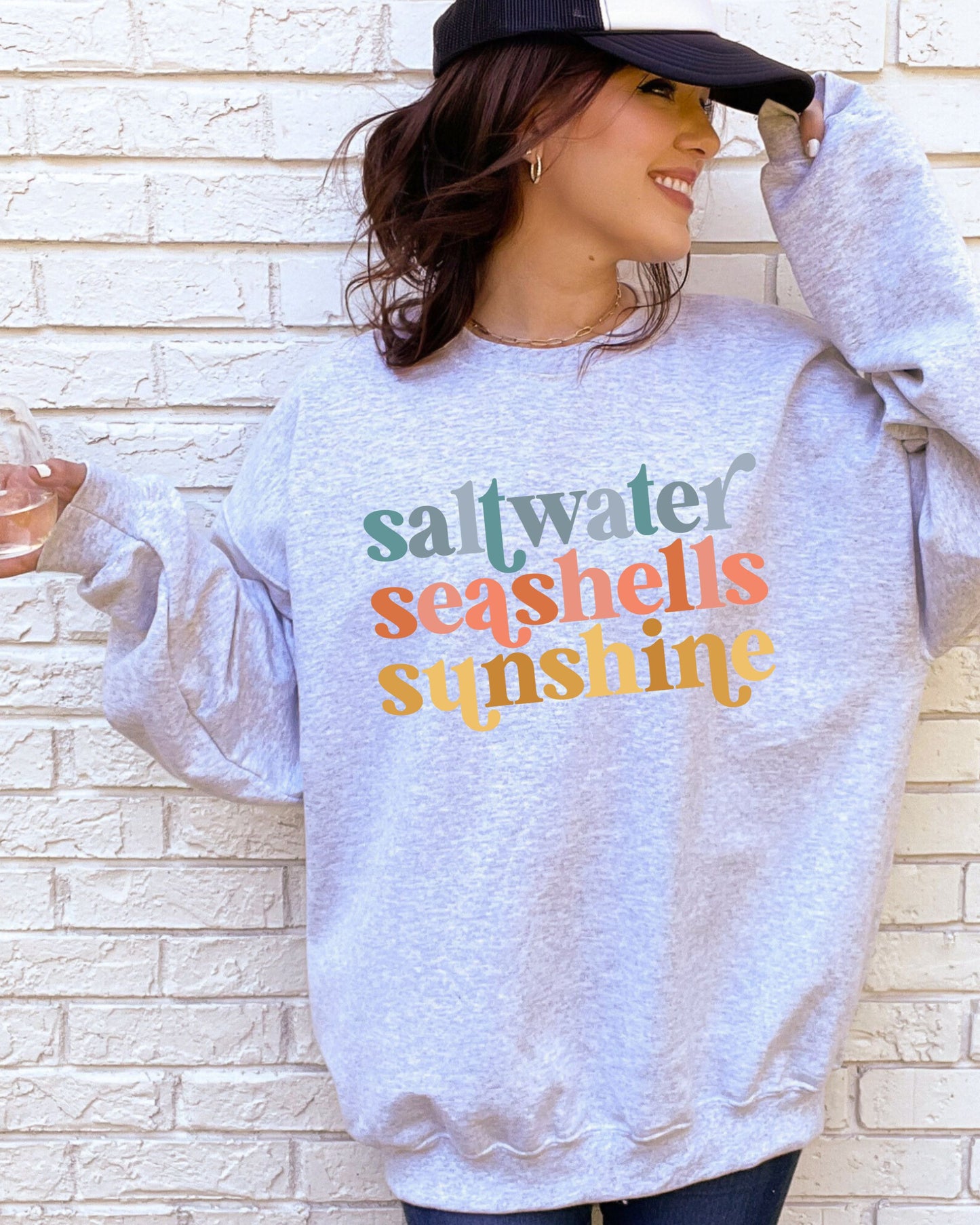 Person wearing a sweatshirt with 'saltwater seashells sunshine' text against a brick wall.