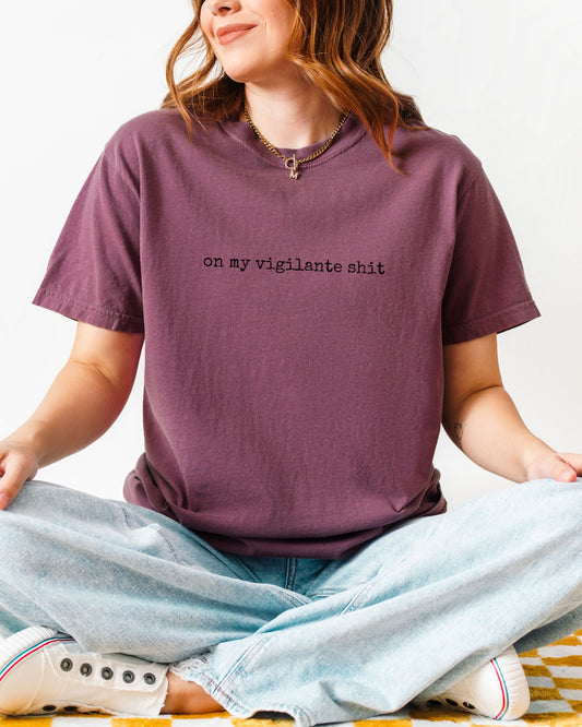 Person wearing a purple t-shirt with on my vigilante shit text in black font sitting on a white background