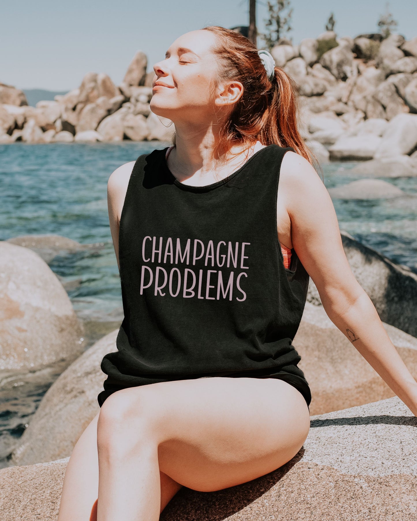 Person wearing a black tank top with 'Champagne Problems' text by a rocky shoreline.