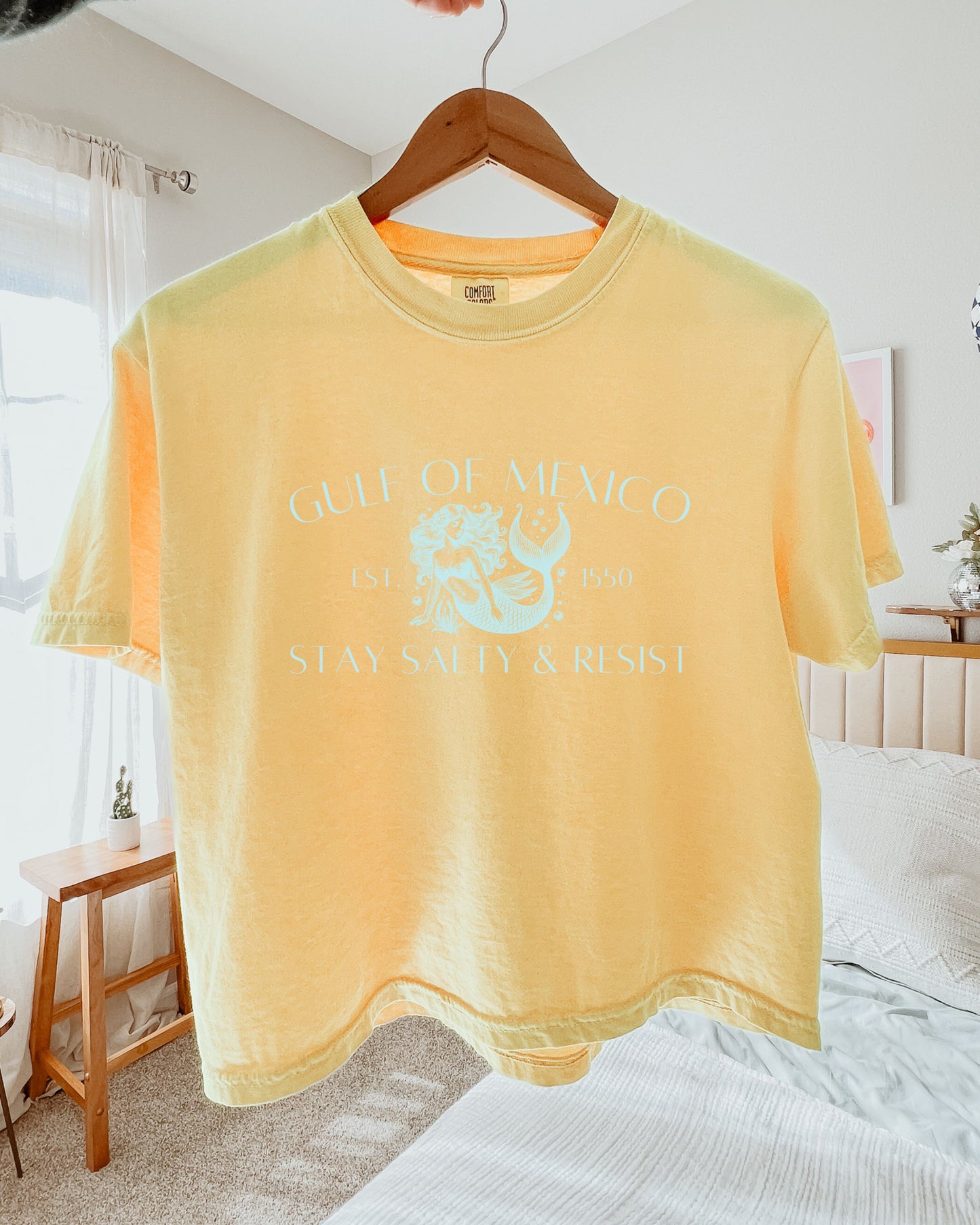 Yellow t-shirt with a logo and text on a hanger in a bedroom setting