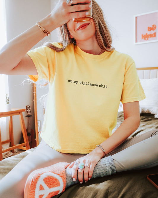 Person wearing a yellow t-shirt with on my vigilante shit text in black font, sitting on a bed in a bright room.
