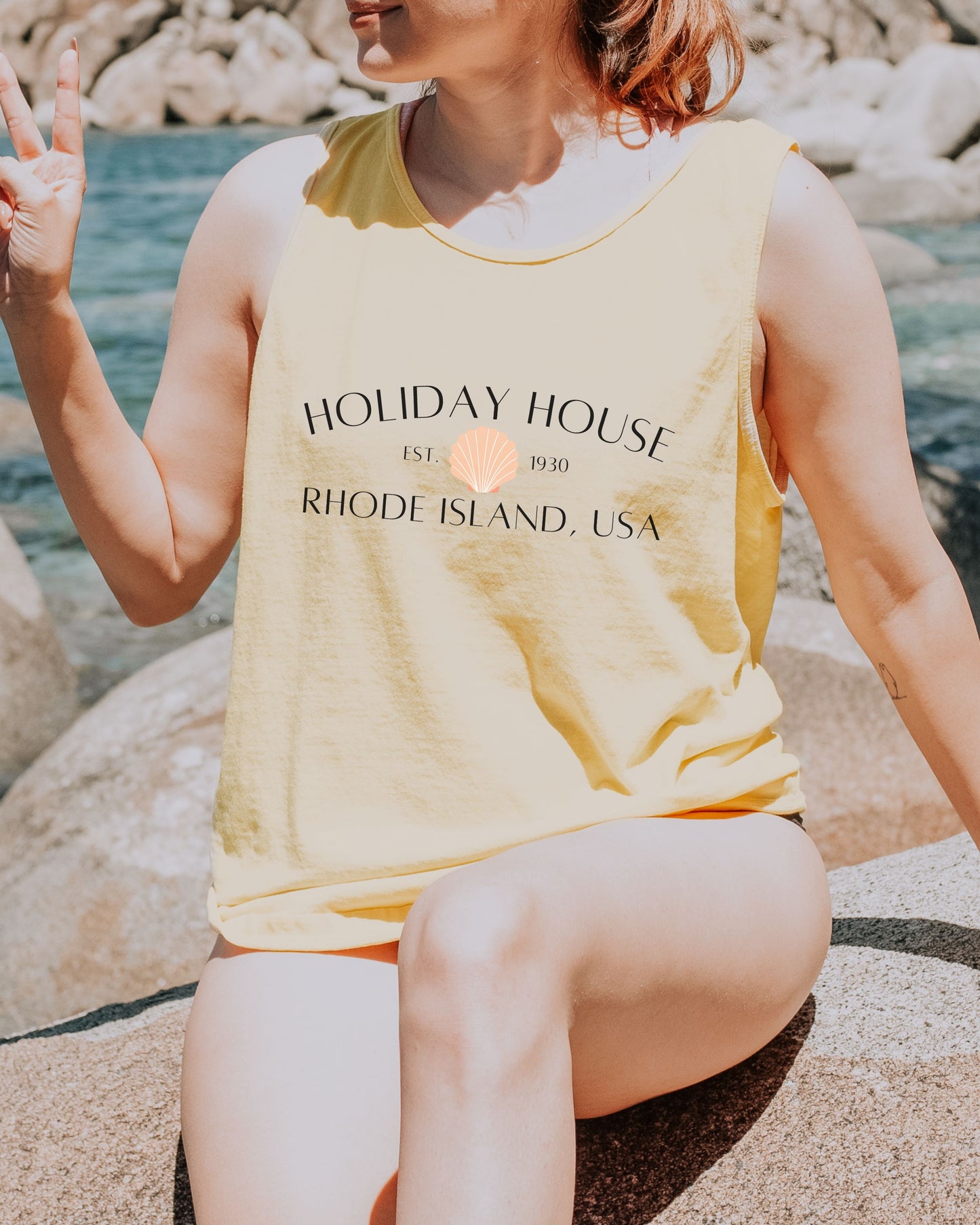Person wearing a yellow tank top with 'Holiday House' text, sitting on rocks by water.