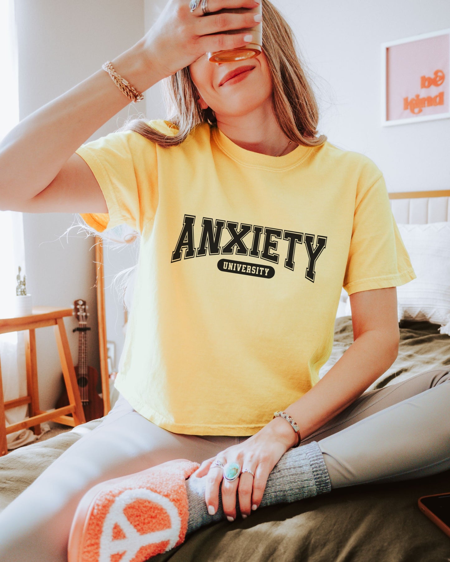 person wearing a yellow tshirt with "Anxiety University" printed on it sitting on a bed