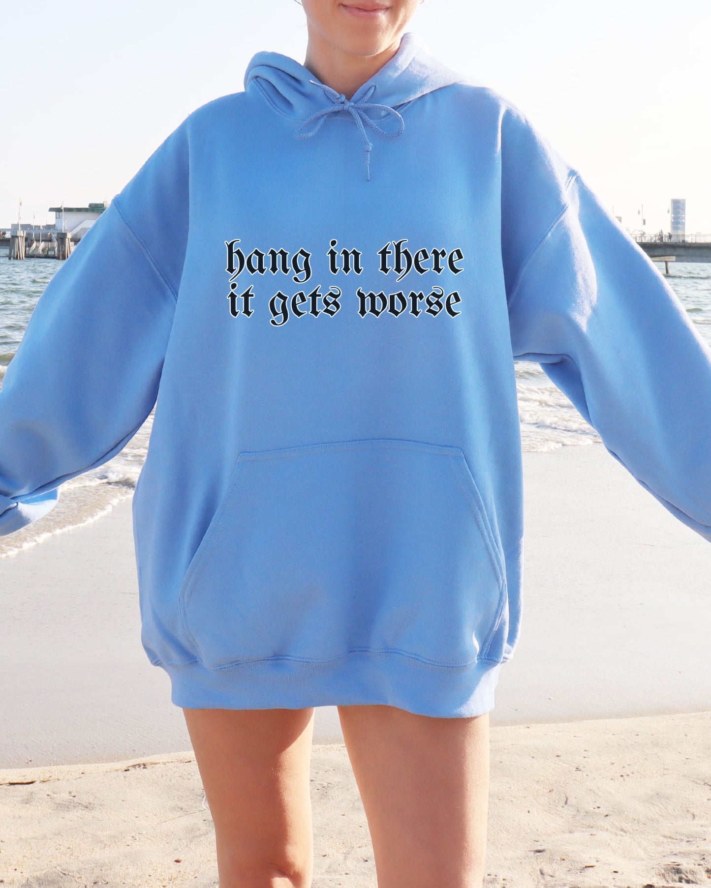 Person wearing a blue hoodie with text 'hang in there it gets worse' by a waterfront.