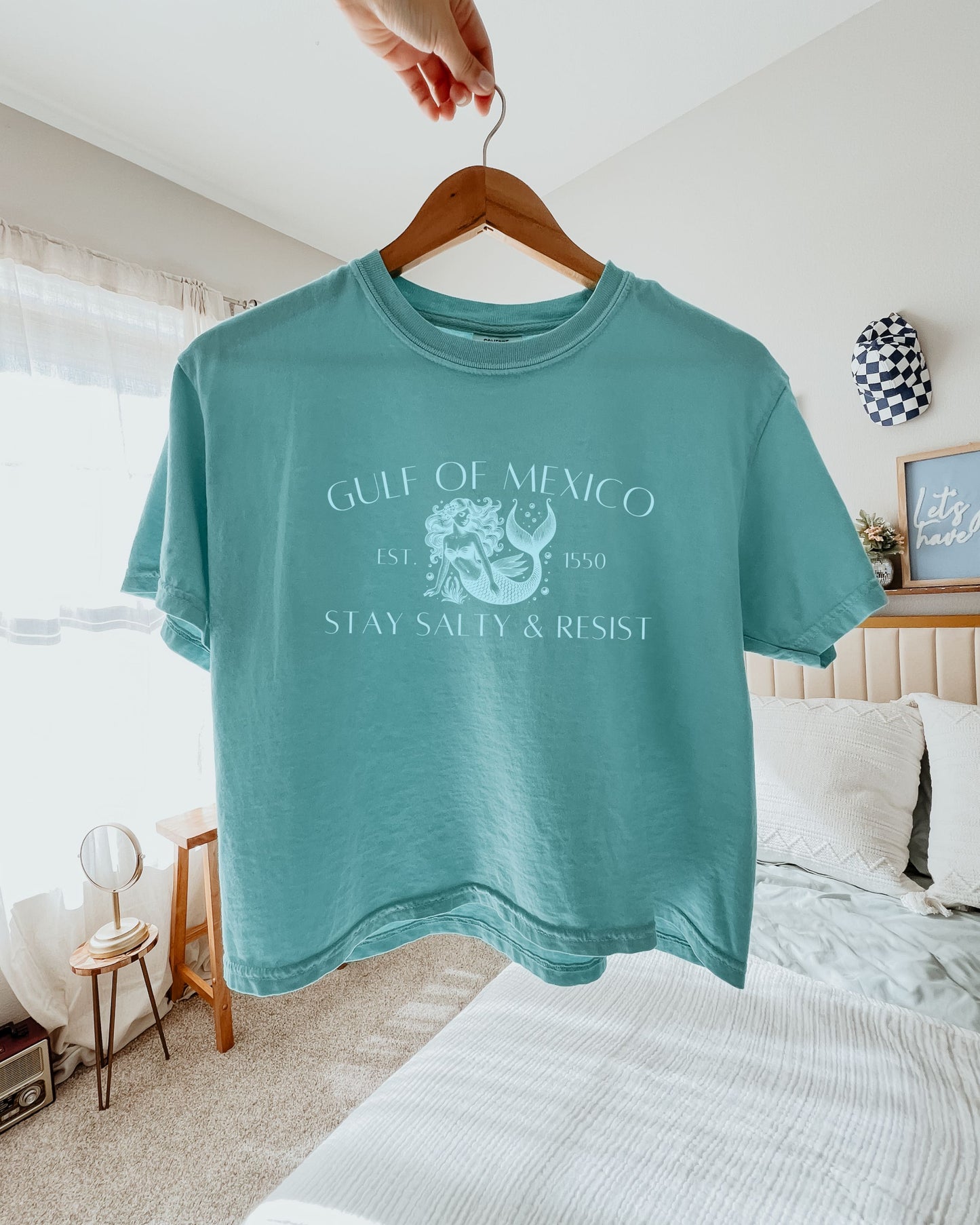 Teal t-shirt with graphic design and text on a hanger in a bedroom setting