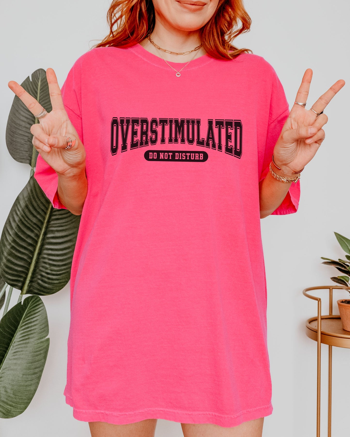 Person wearing a bright pink oversized t-shirt with 'OVERSTIMULATED DO NOT DISTURB' text, making peace signs.