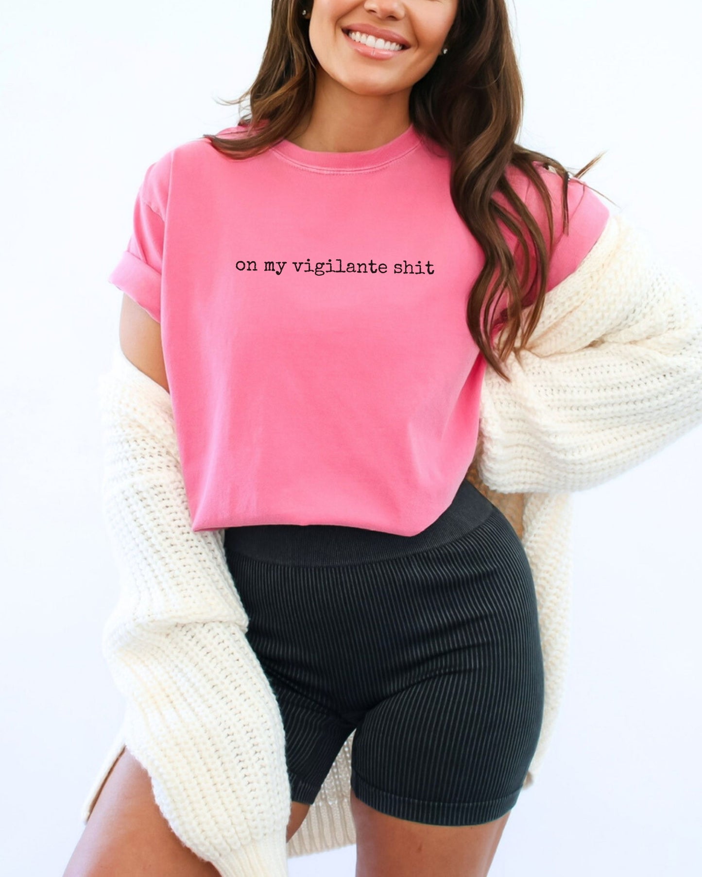Person wearing a pink shirt with on my vigilante shit text in black font and black shorts on a white background