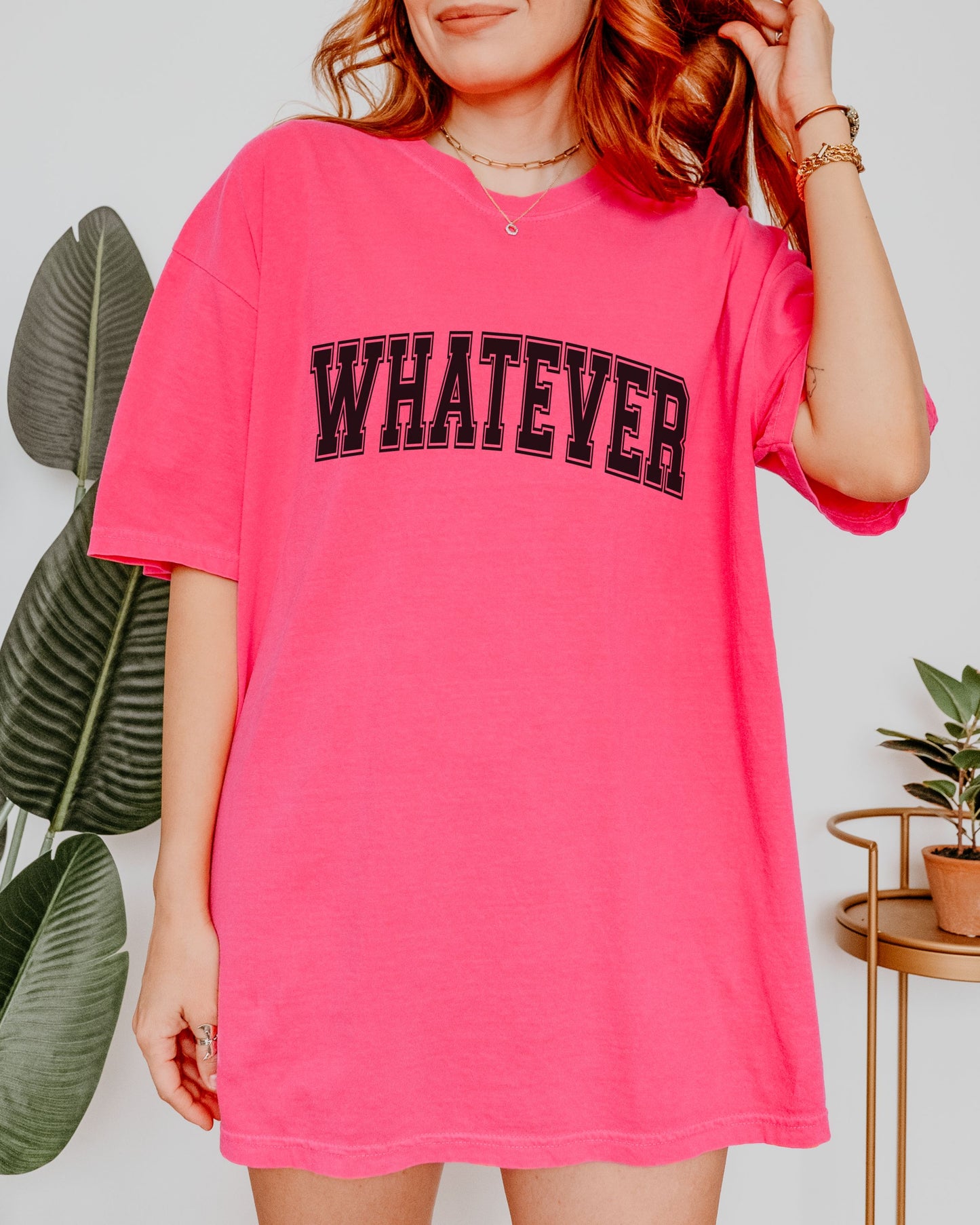 Person wearing a bright pink t-shirt with 'WHATEVER' text, standing indoors with plants in the background.