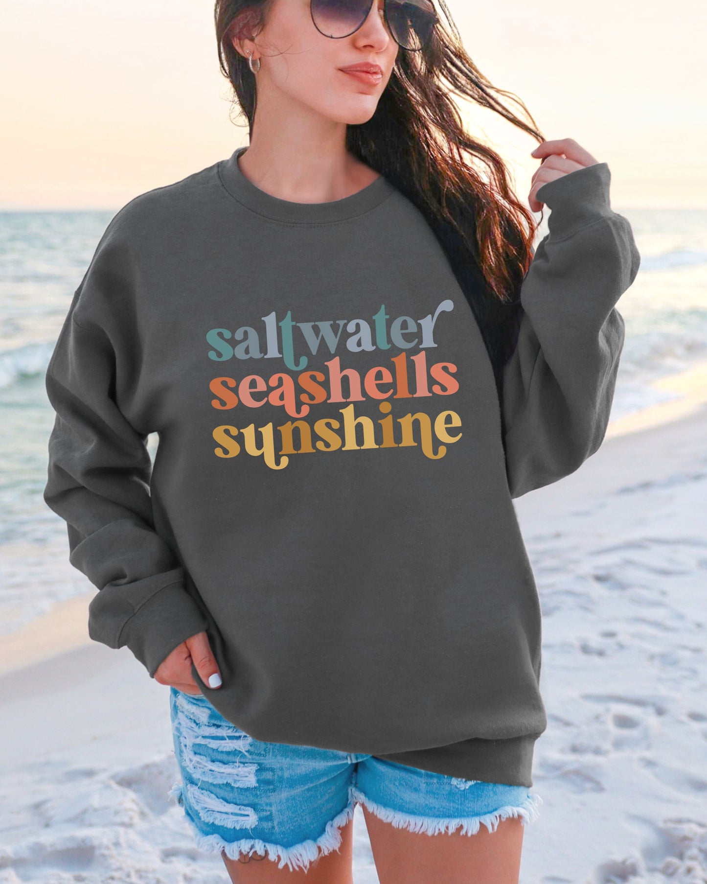 Person wearing a sweatshirt with 'saltwater seashells sunshine' text on a beach.