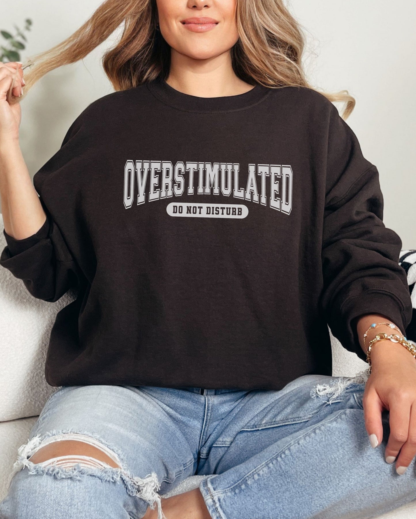 Person wearing a black sweatshirt with 'OVERSTIMULATED DO NOT DISTURB' text, sitting on a white couch.