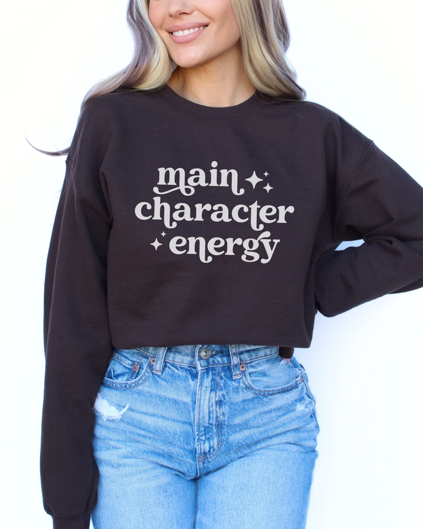 Person wearing a chocolate brown sweatshirt with 'main character energy' text and blue jeans.