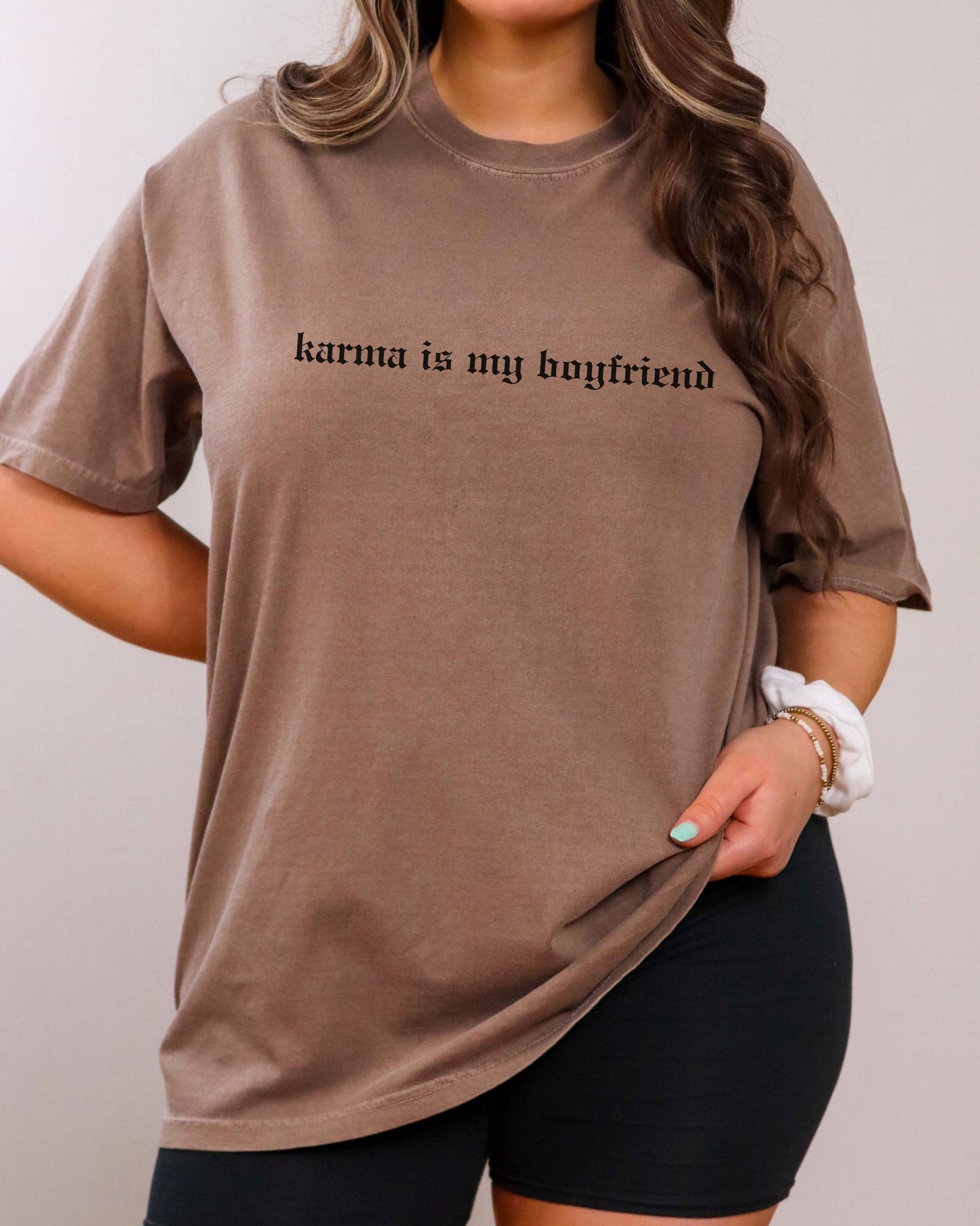 Person wearing a brown t-shirt with 'karma is my boyfriend' text on a plain background