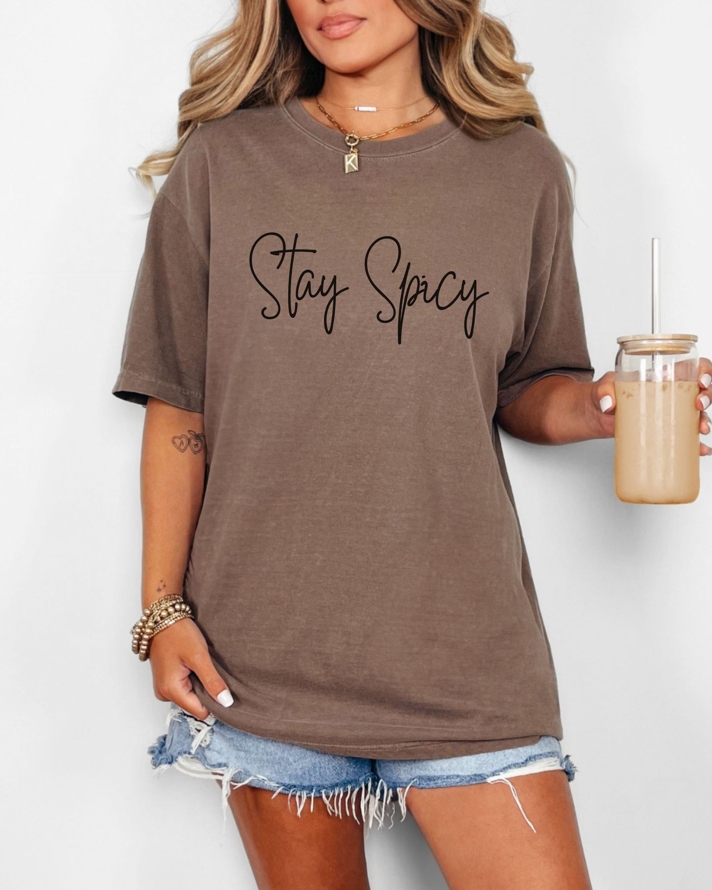Woman wearing a brown t-shirt with 'Stay Spicy' text, holding a drink.