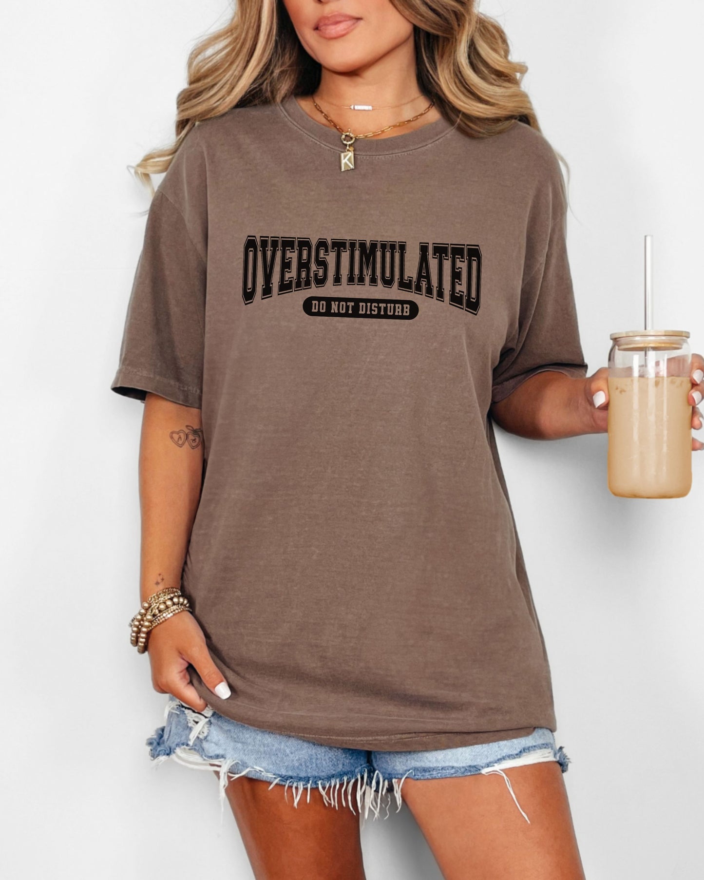 Person wearing a brown t-shirt with 'OVERSTIMULATED DO NOT DISTURB' text, holding a drink.