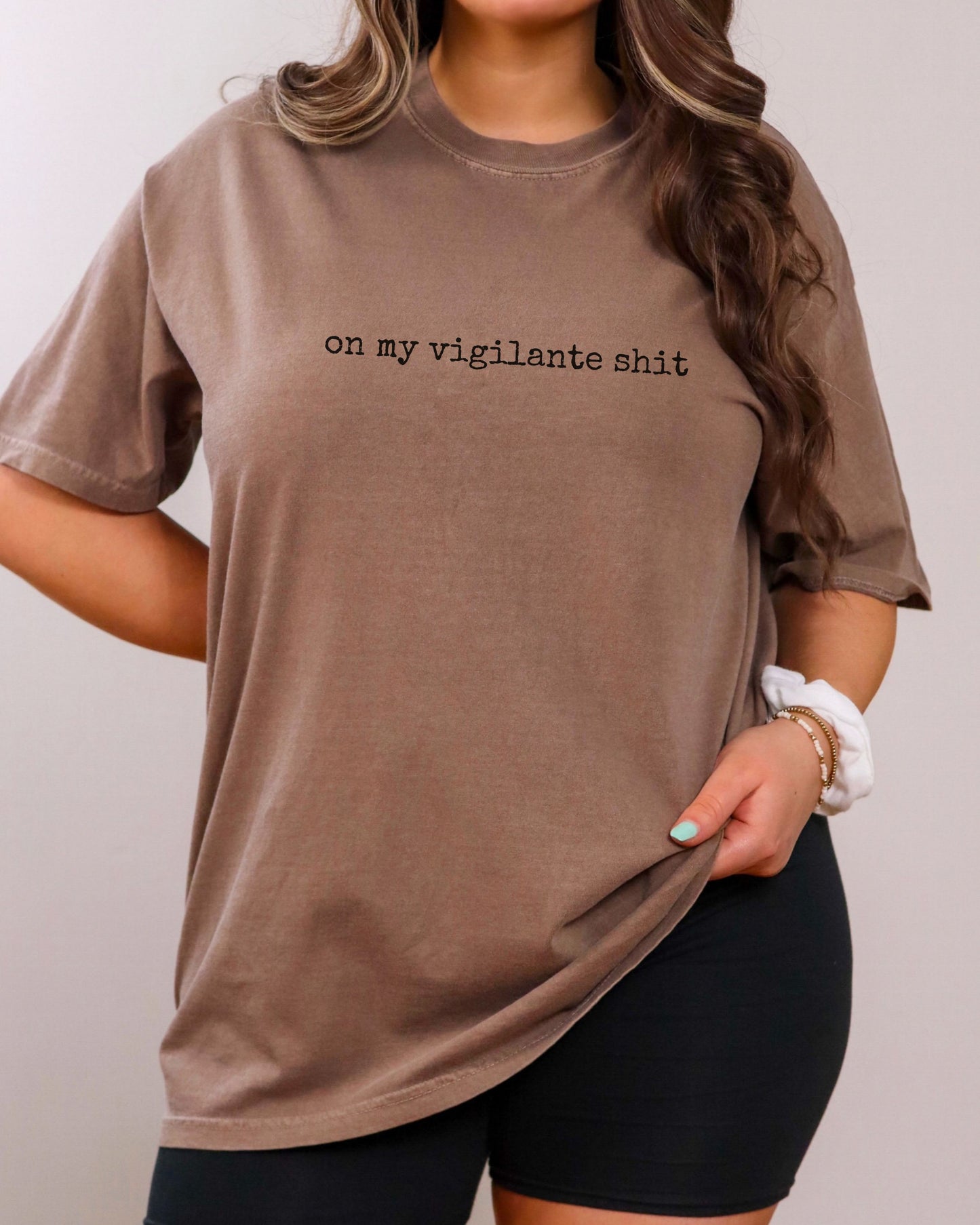 Person wearing a brown t-shirt with on my vigilante shit text in black font on a plain background