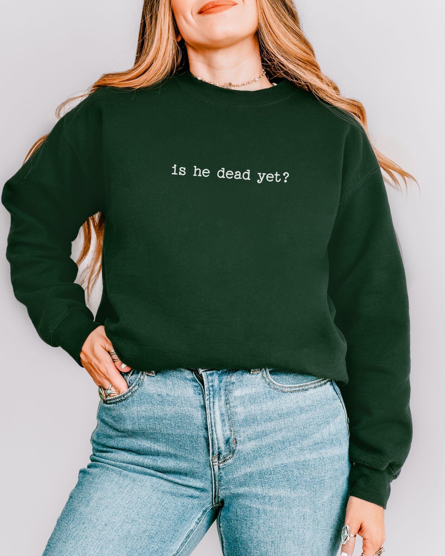 person wearing a forest green crewneck sweatshirt with "is he dead yet?" printed in a typewriter font standing in front of a white wall