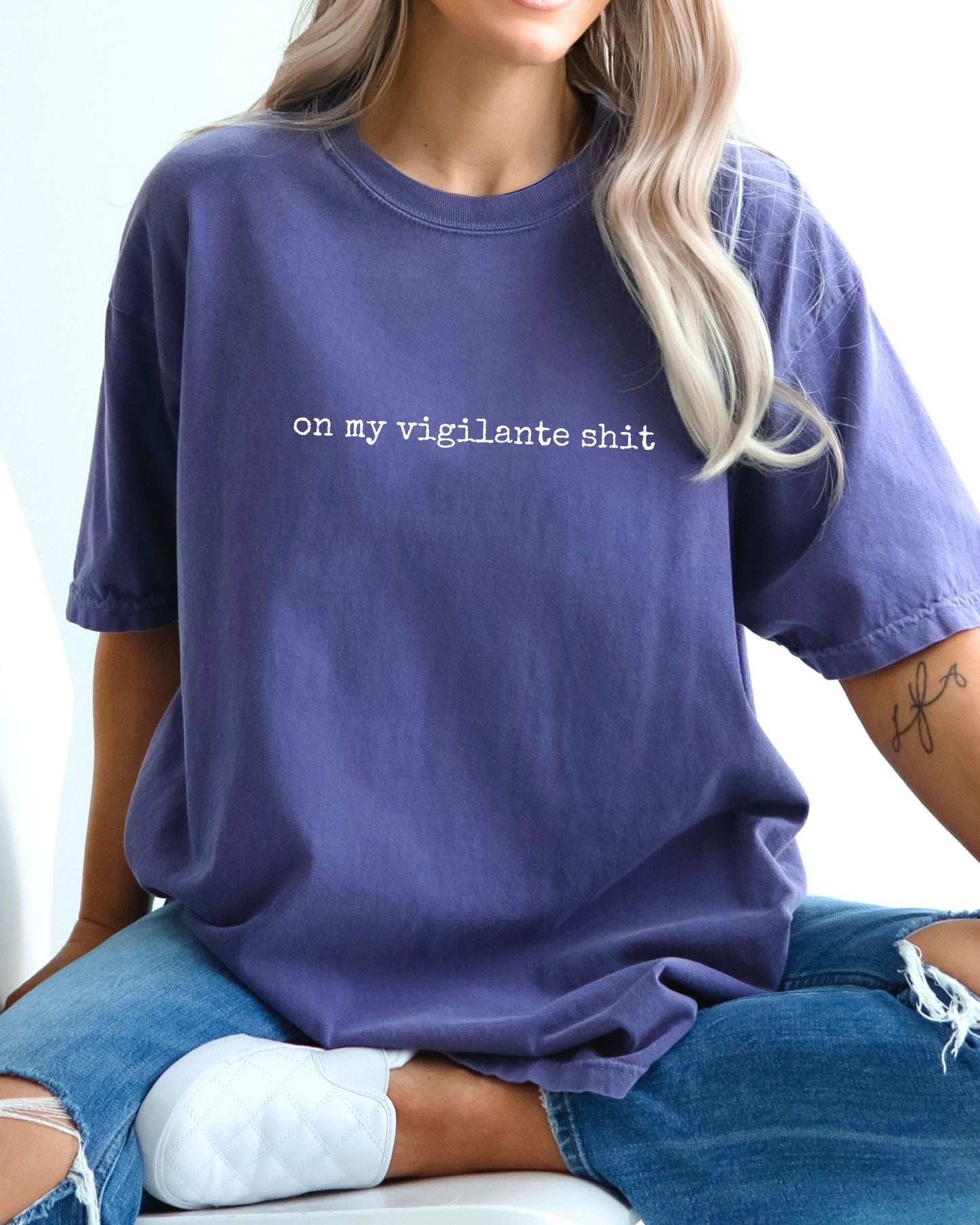Person wearing a purple t-shirt with on my vigilante shit text in white font, sitting on a white surface.