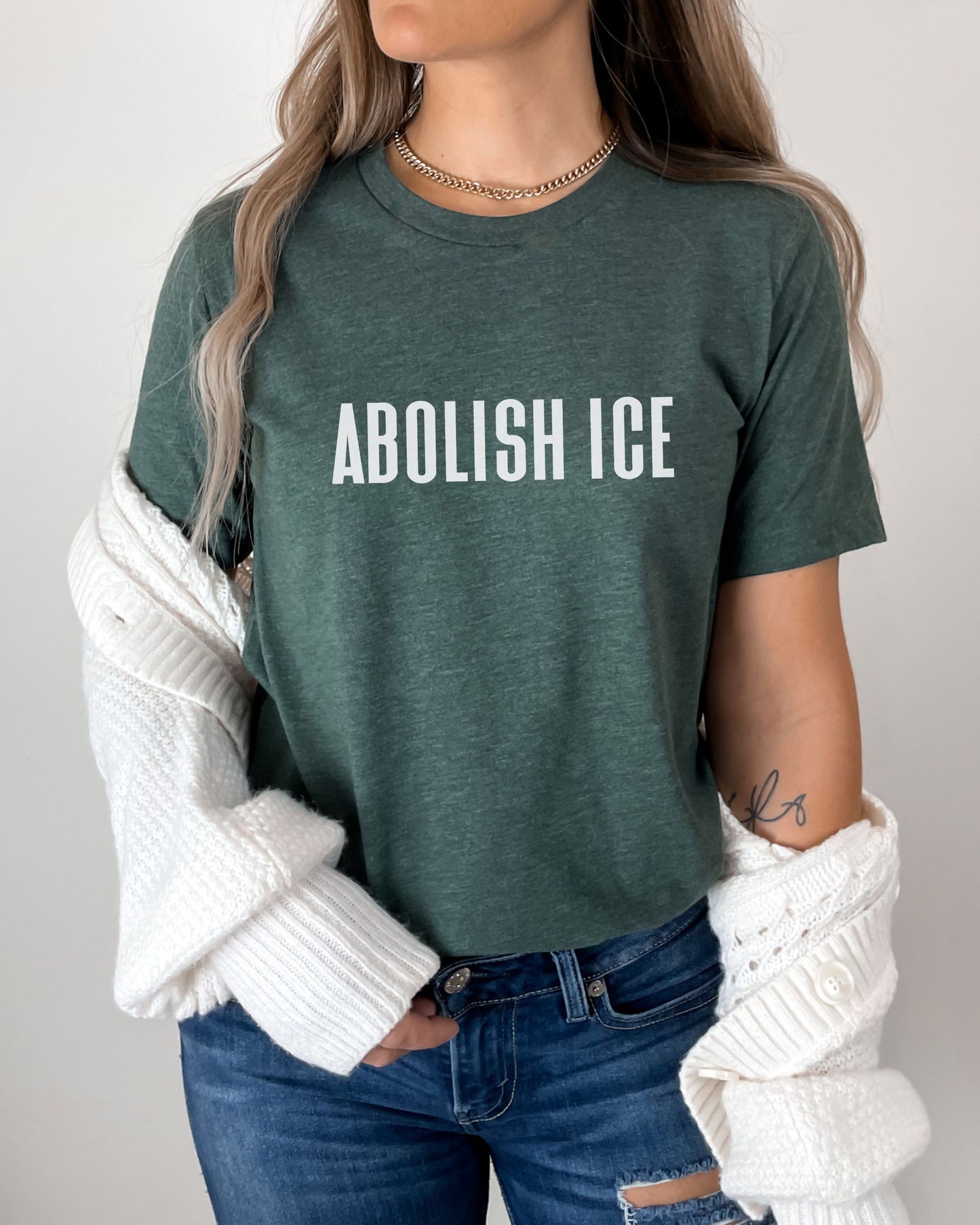Person wearing a green t-shirt with 'ABOLISH ICE' text, white sweater, and blue jeans.