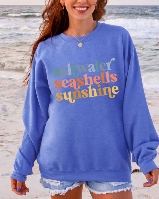 Woman wearing a blue sweatshirt with 'saltwater seashells sunshine' text on a beach.