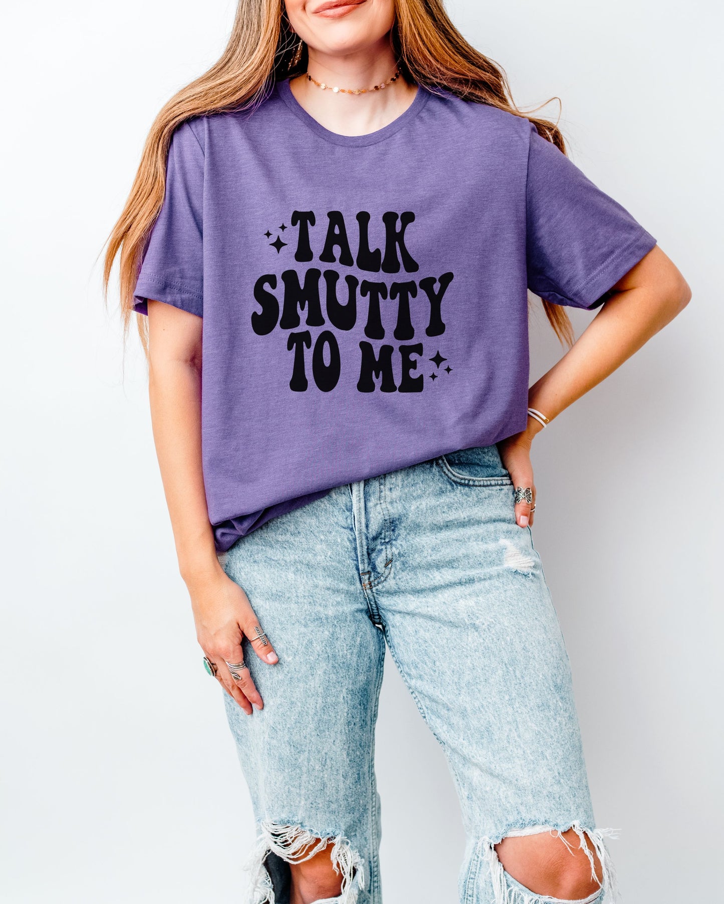 Person wearing a purple t-shirt with 'Talk Smutty To Me' text and ripped jeans on a plain background