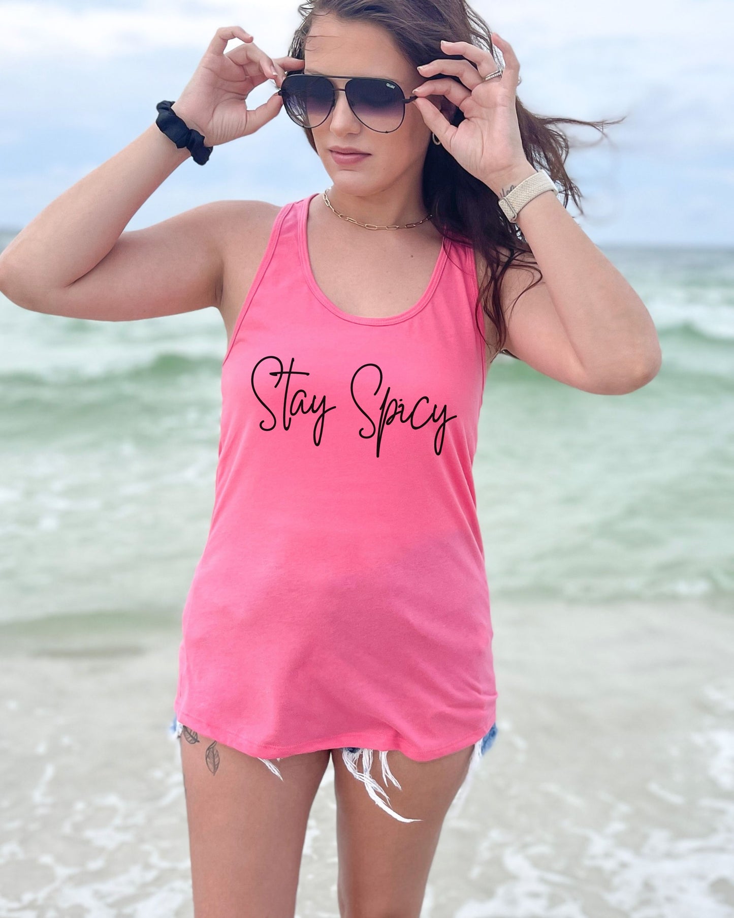 Woman wearing a pink tank top with 'Stay Spicy' text on a beach.