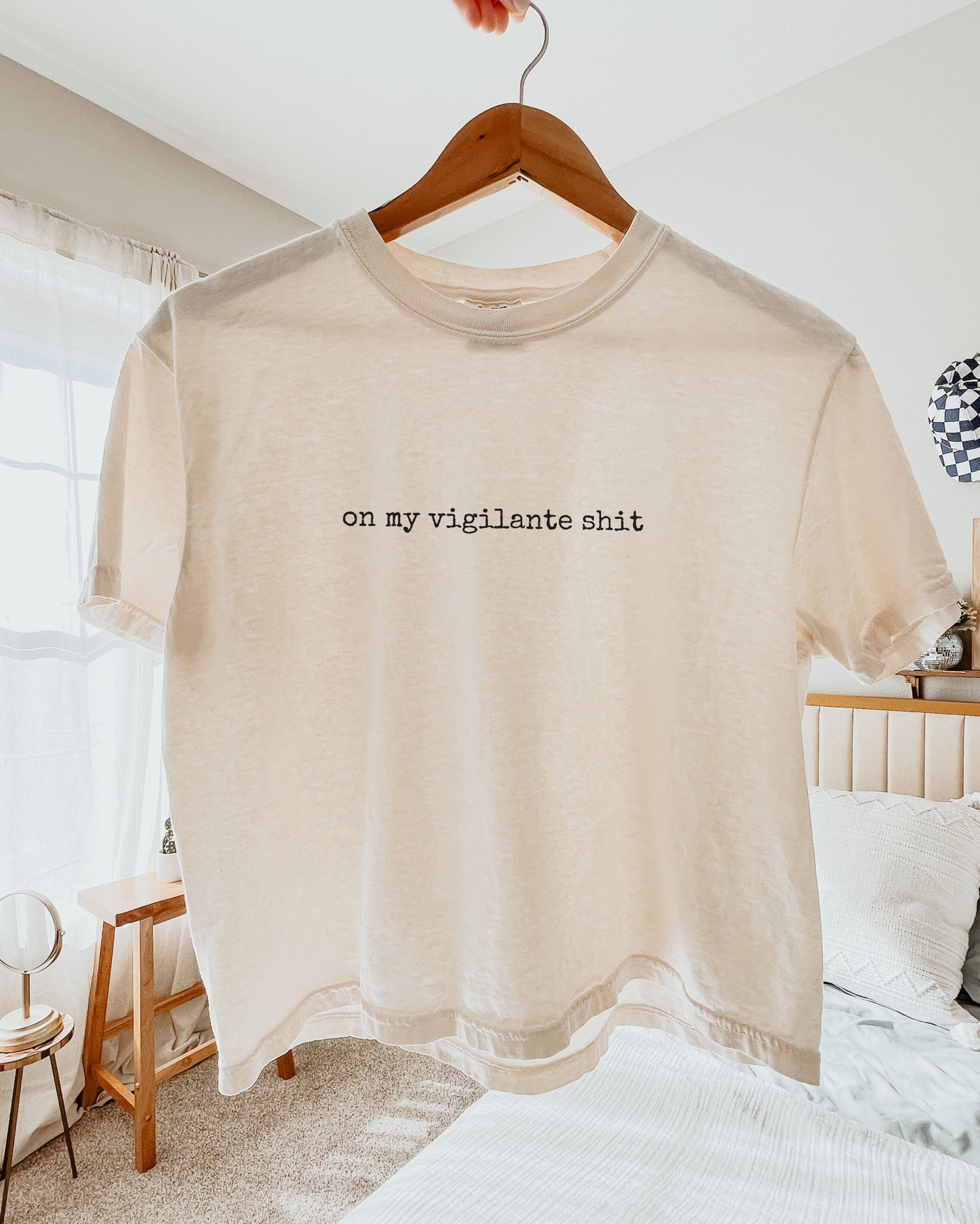 Beige t-shirt with on my vigilante shit text in black font on a hanger in a bedroom