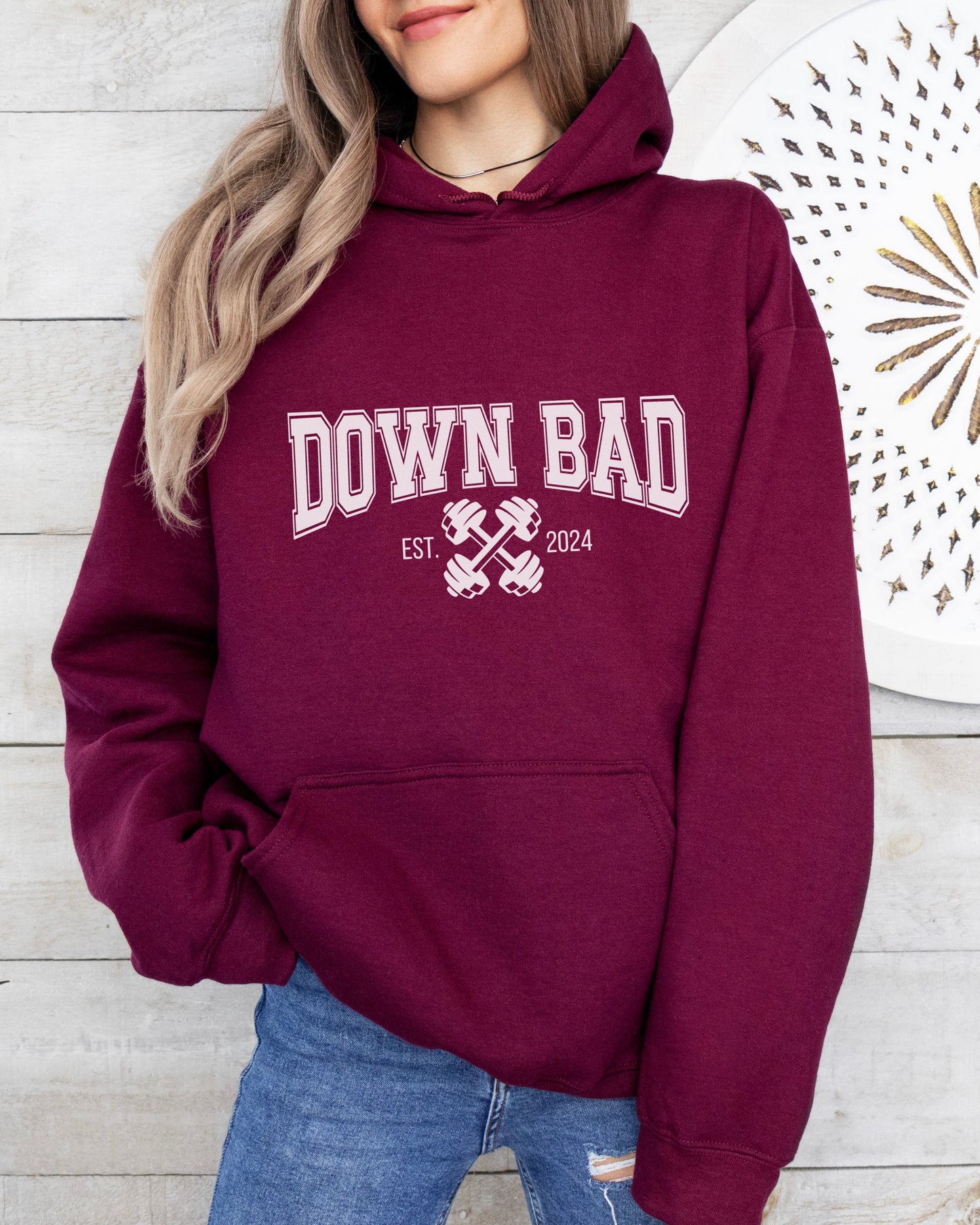 Maroon hoodie with 'DOWN BAD' text worn by a person against a wooden wall.