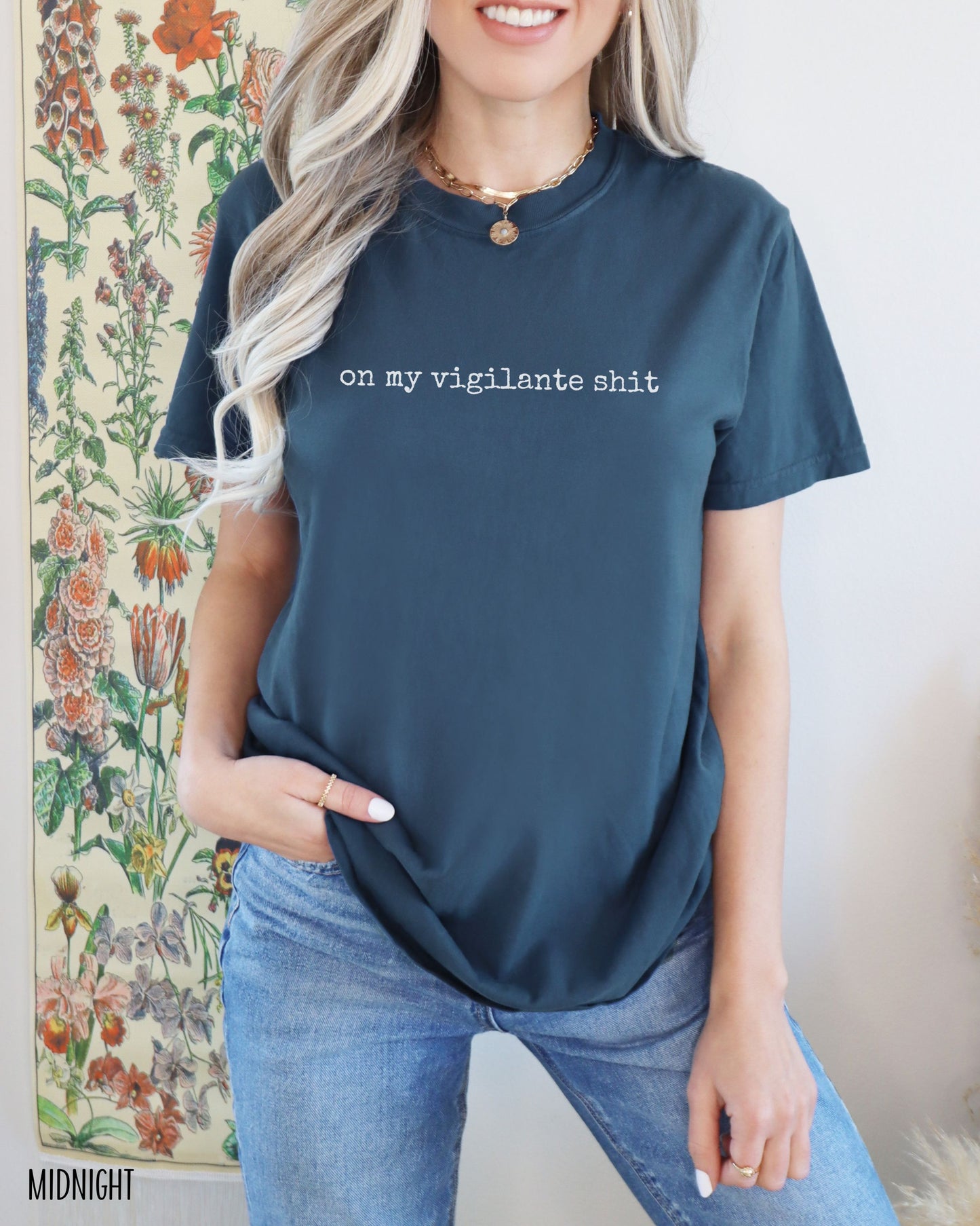 Person wearing a blue t-shirt with on my vigilante shit text in white font, standing in front of a floral wall.