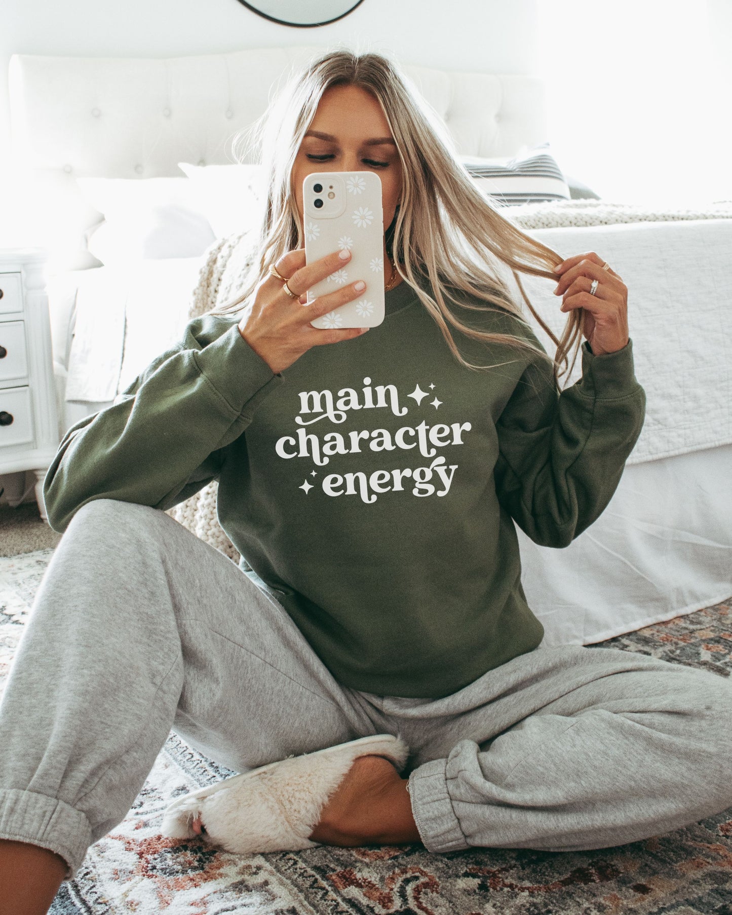 Person wearing a green sweatshirt with 'main character energy' text, sitting on a bed.