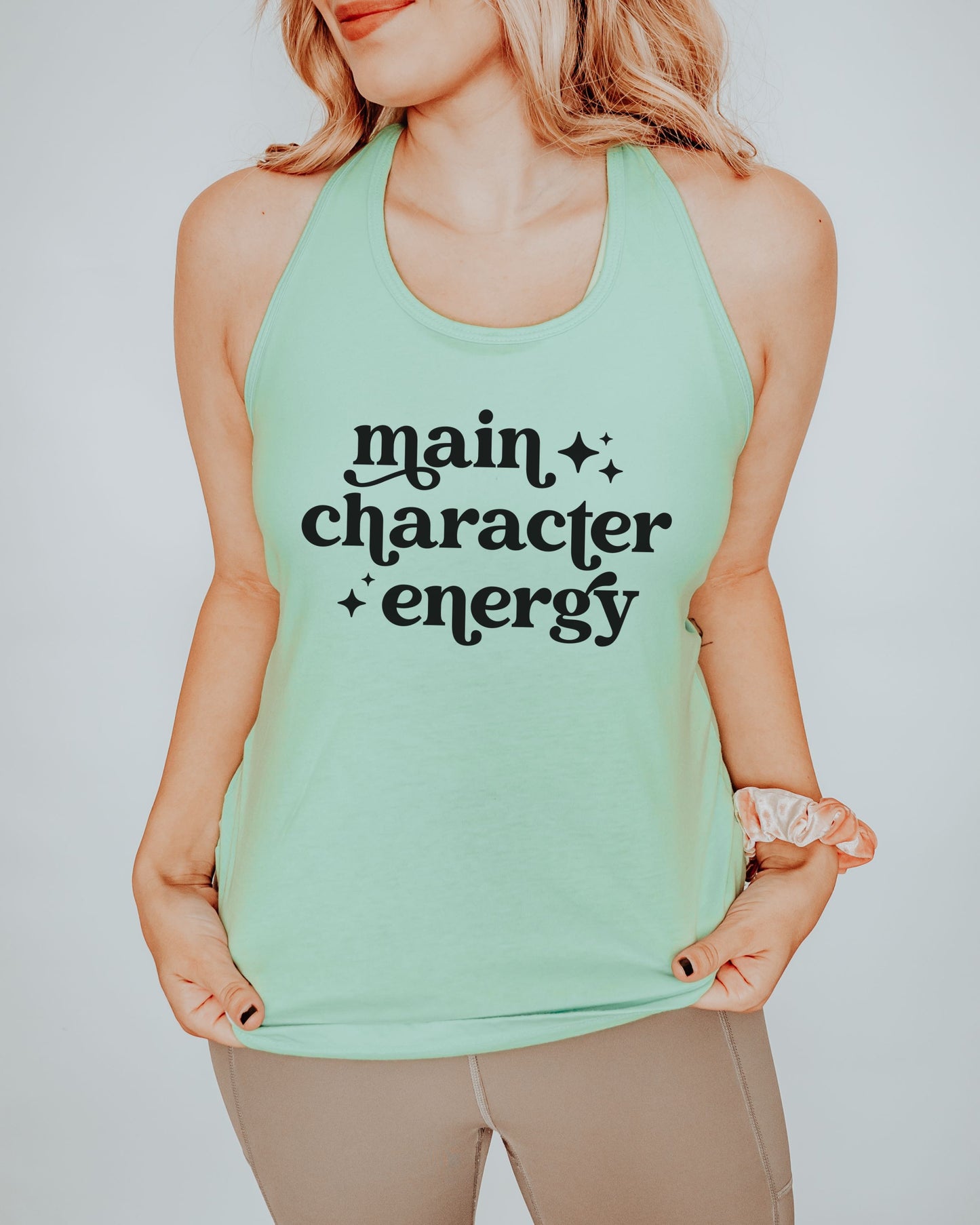 Person wearing a mint green tank top with 'main character energy' text on a plain background