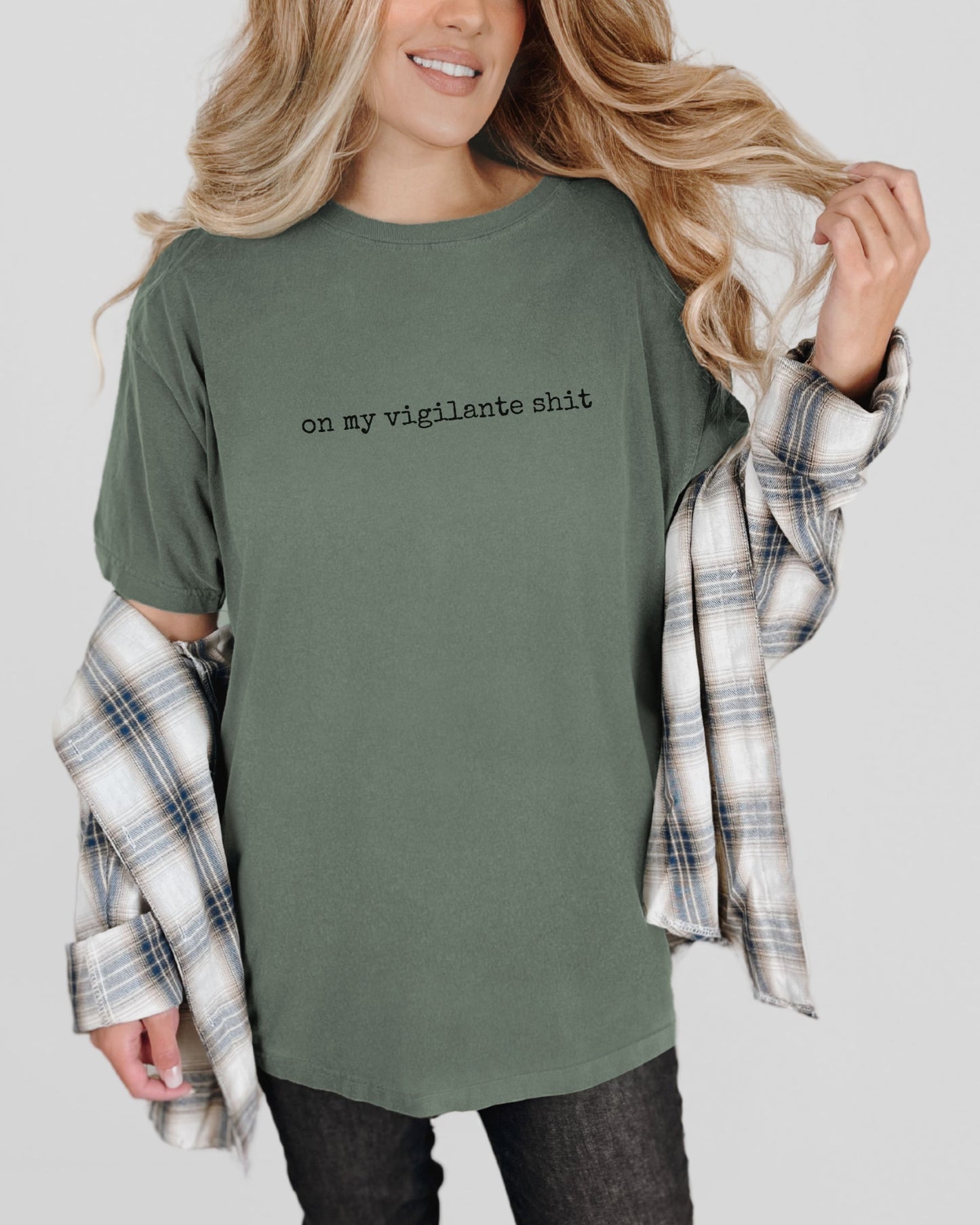 Person wearing a green t-shirt with on my vigilante shit text with black font and plaid shirt over shoulders on a plain background