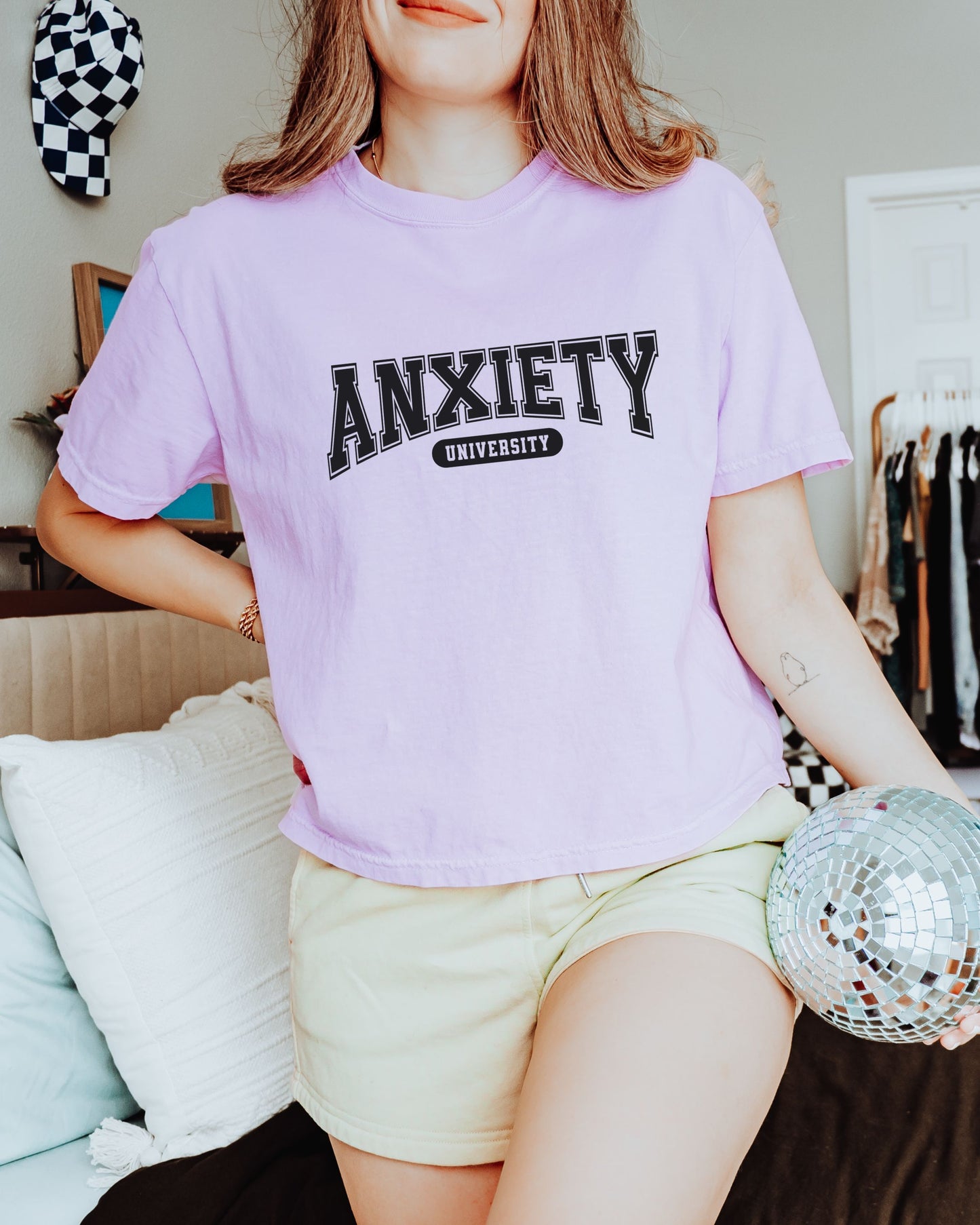 Person wearing a light purple t-shirt with 'ANXIETY UNIVERSITY' text, holding a disco ball indoors.