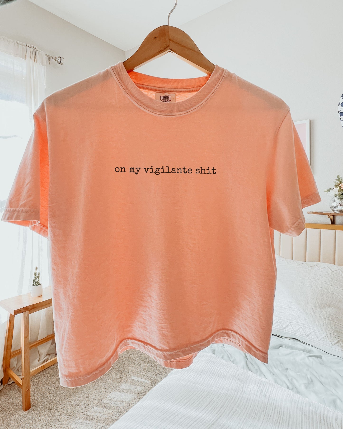 Peach t-shirt with on my vigilante shit text in black font on a hanger in a bedroom