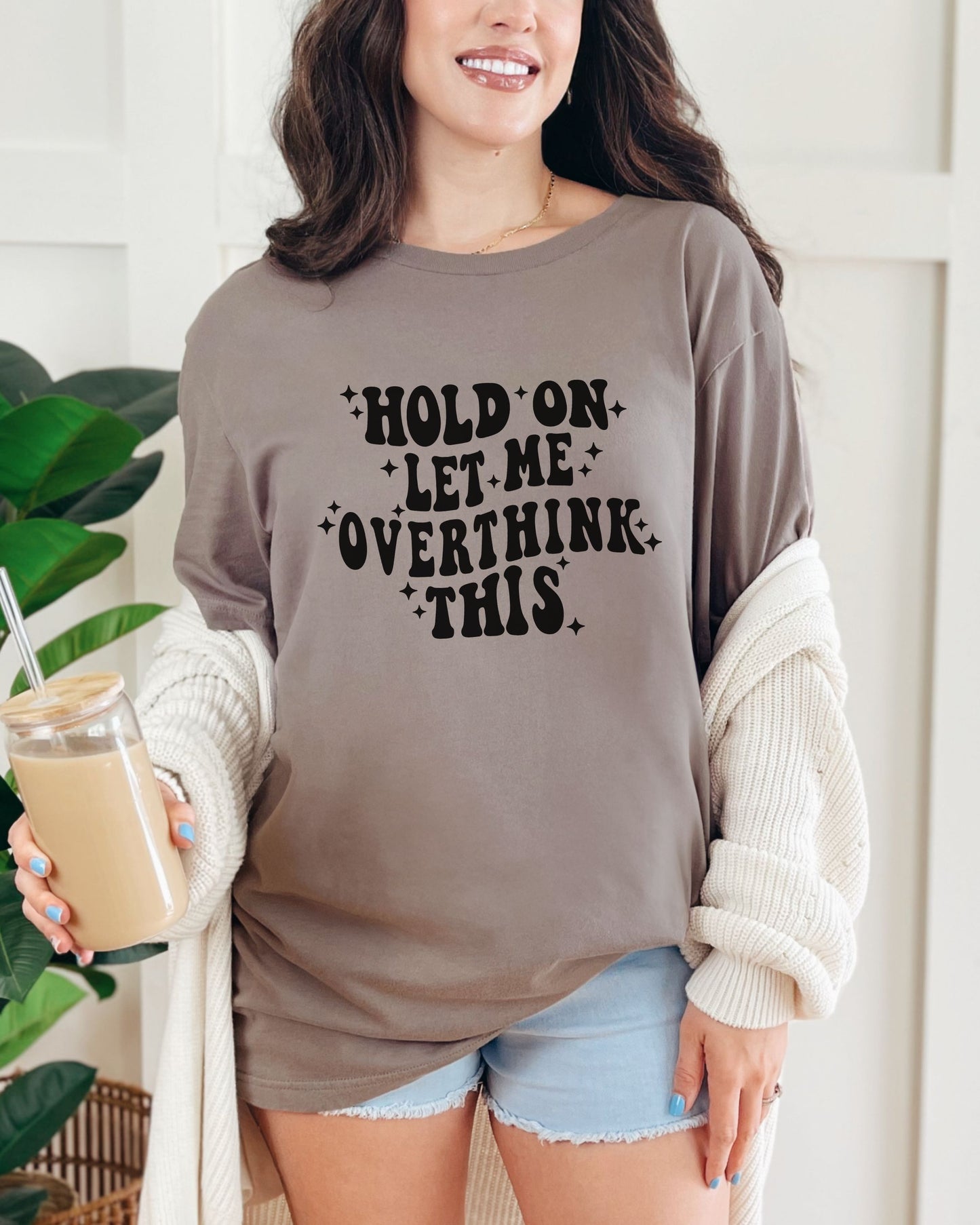 Woman wearing a brown tshirt with text, holding a drink, standing indoors.