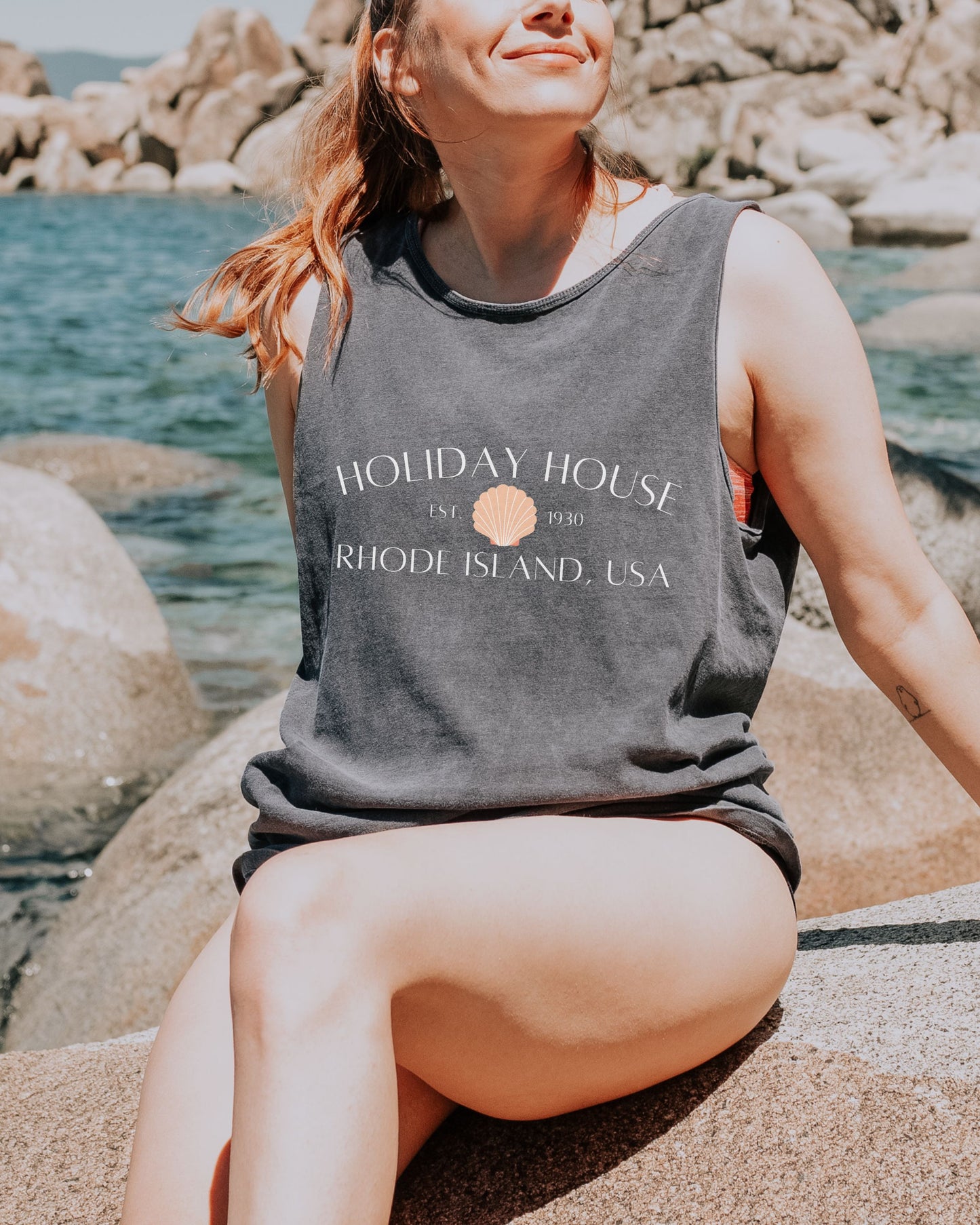 Woman wearing a tank top with 'Holiday House' text sitting on rocks by the water.