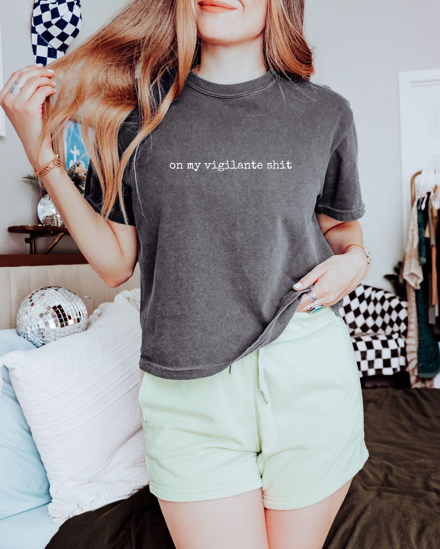 Person wearing a dark gray t-shirt with on my vigilante shit text in white font and light green shorts in a casual setting.