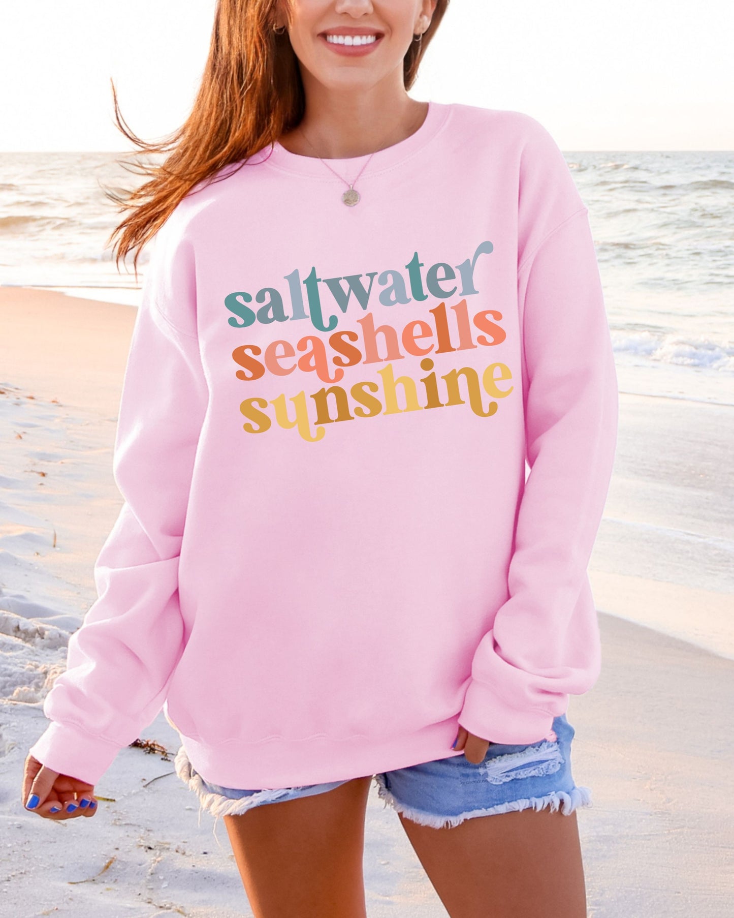 Person wearing a pink sweatshirt with 'saltwater seashells sunshine' text on a beach.
