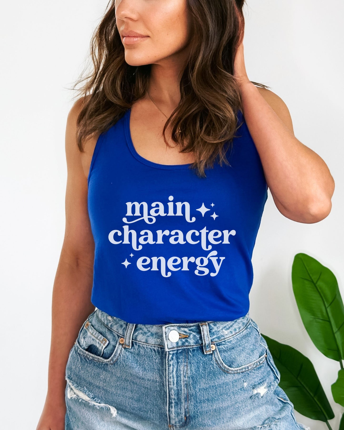 Person wearing a blue tank top with 'main character energy' text, standing against a white background with a plant.
