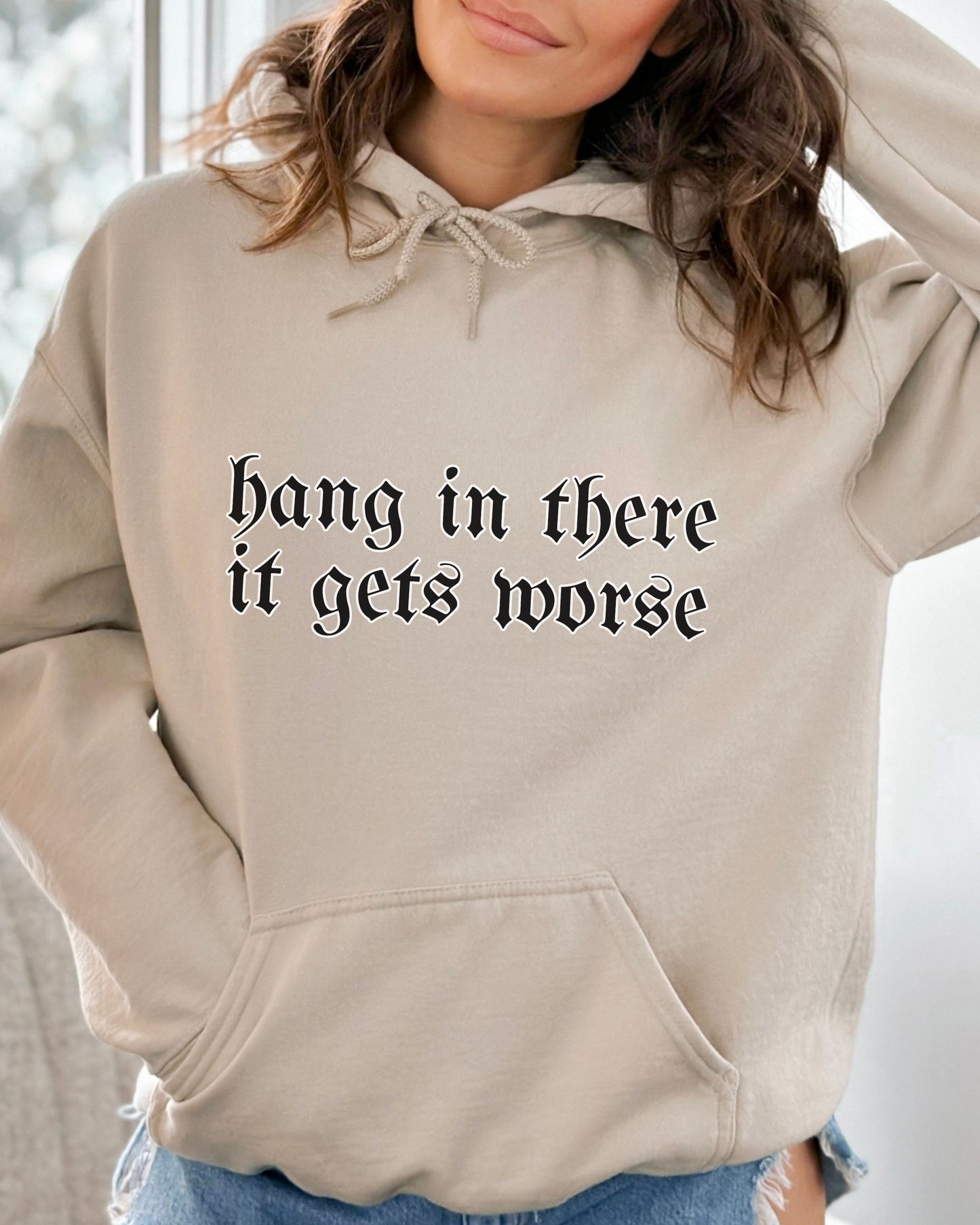 Beige hoodie with text 'hang in there it gets worse' worn by a person.