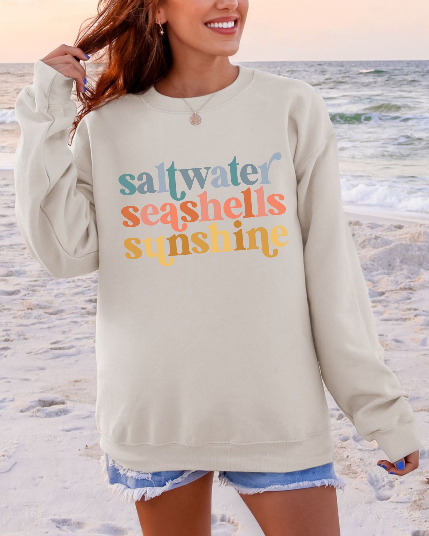 Person wearing a beige sweatshirt with 'saltwater seashells sunshine' text on a beach.