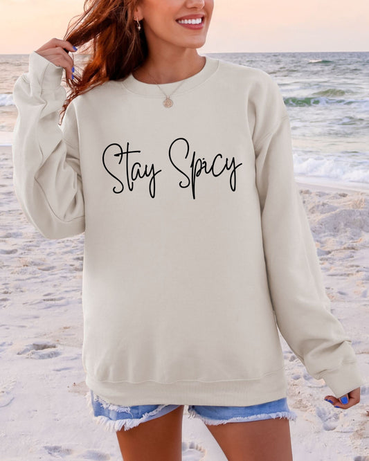 Person wearing a beige sweatshirt with 'Stay Spicy' text on a beach.
