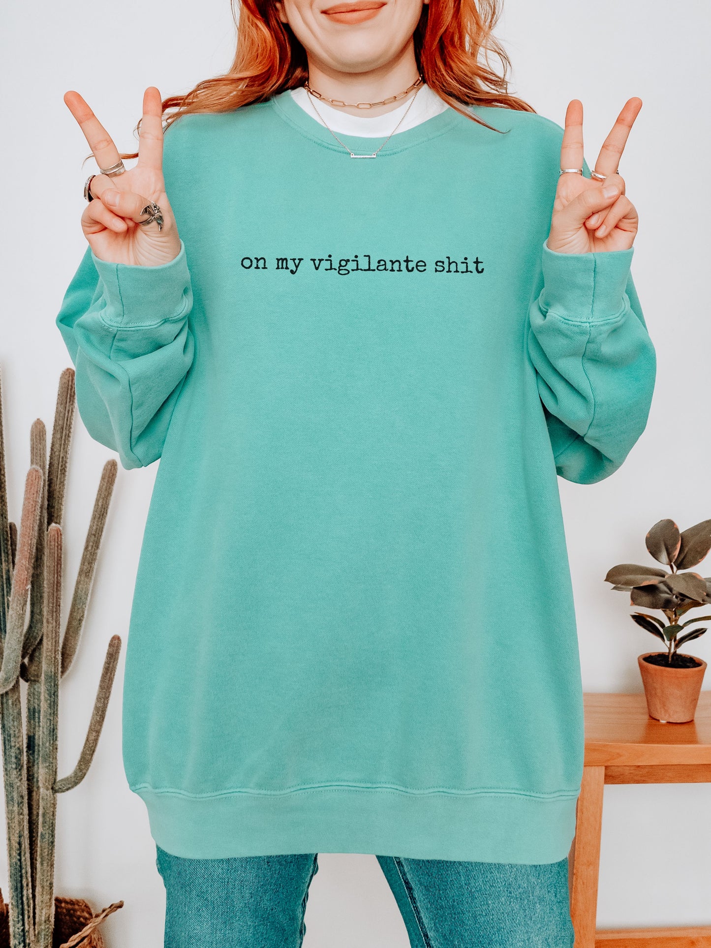 Person wearing a teal sweatshirt with text, making peace signs, in a room with plants.