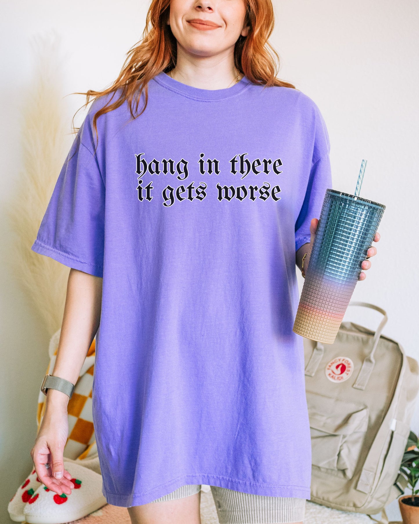 Person wearing a purple t-shirt with text, holding a blue tumbler, with a neutral background