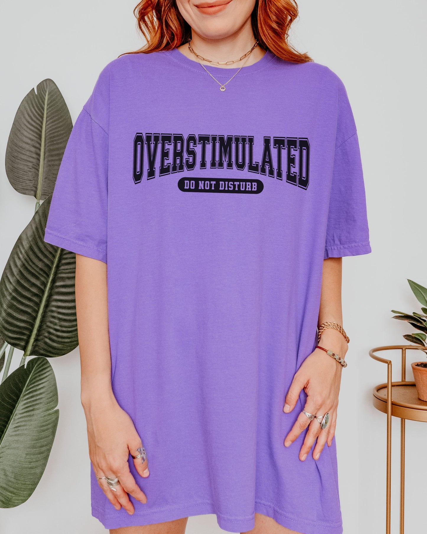 Person wearing a purple t-shirt with 'OVERSTIMULATED DO NOT DISTURB' text, standing indoors with plants in the background.