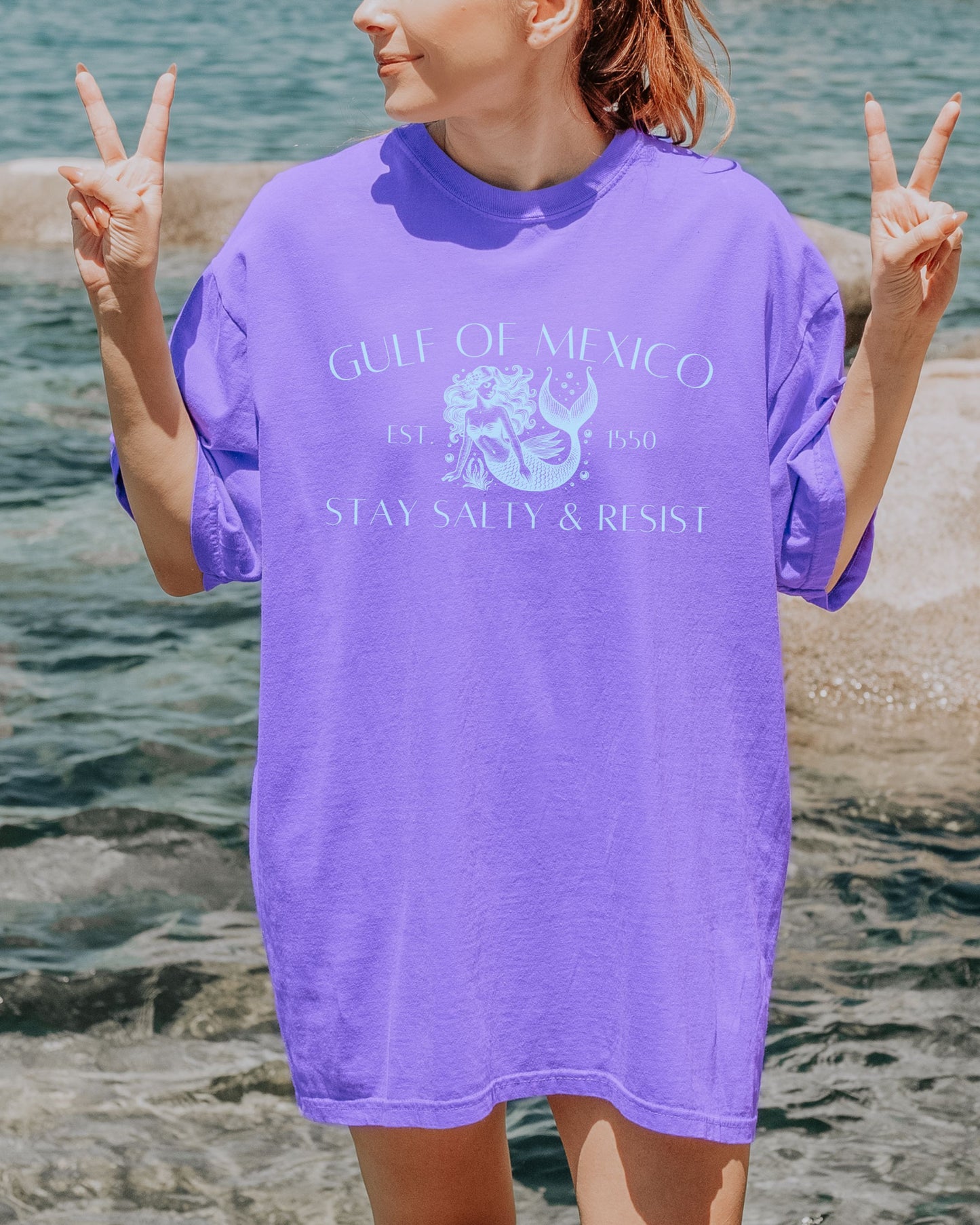 Person wearing a purple t-shirt with 'Gulf of Mexico' text and graphics, standing by water.